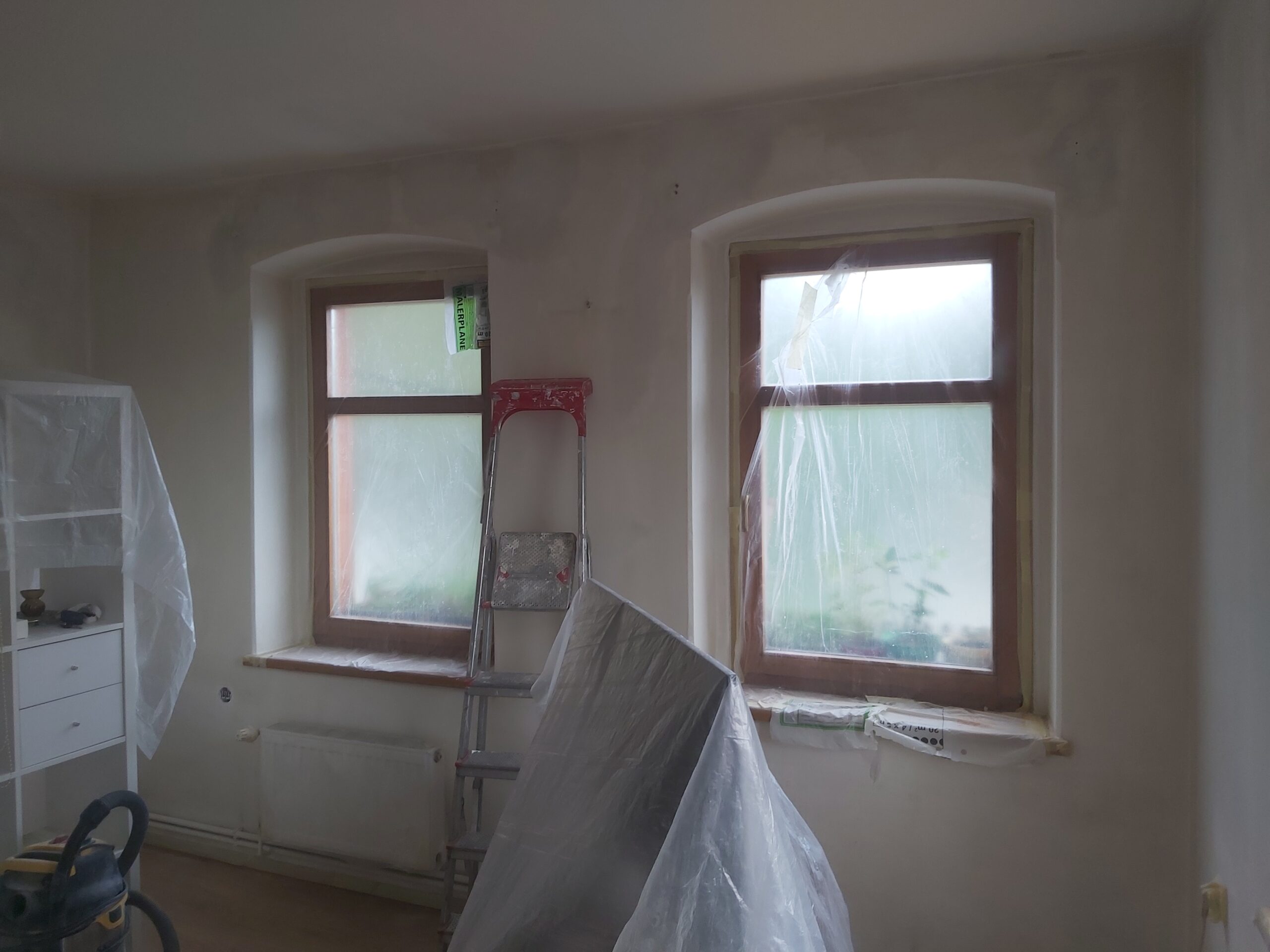 Dwa okna w remontowanym pokoju, osłonięte folią ochronną, z drabiną i meblami zabezpieczonymi przed zabrudzeniem.