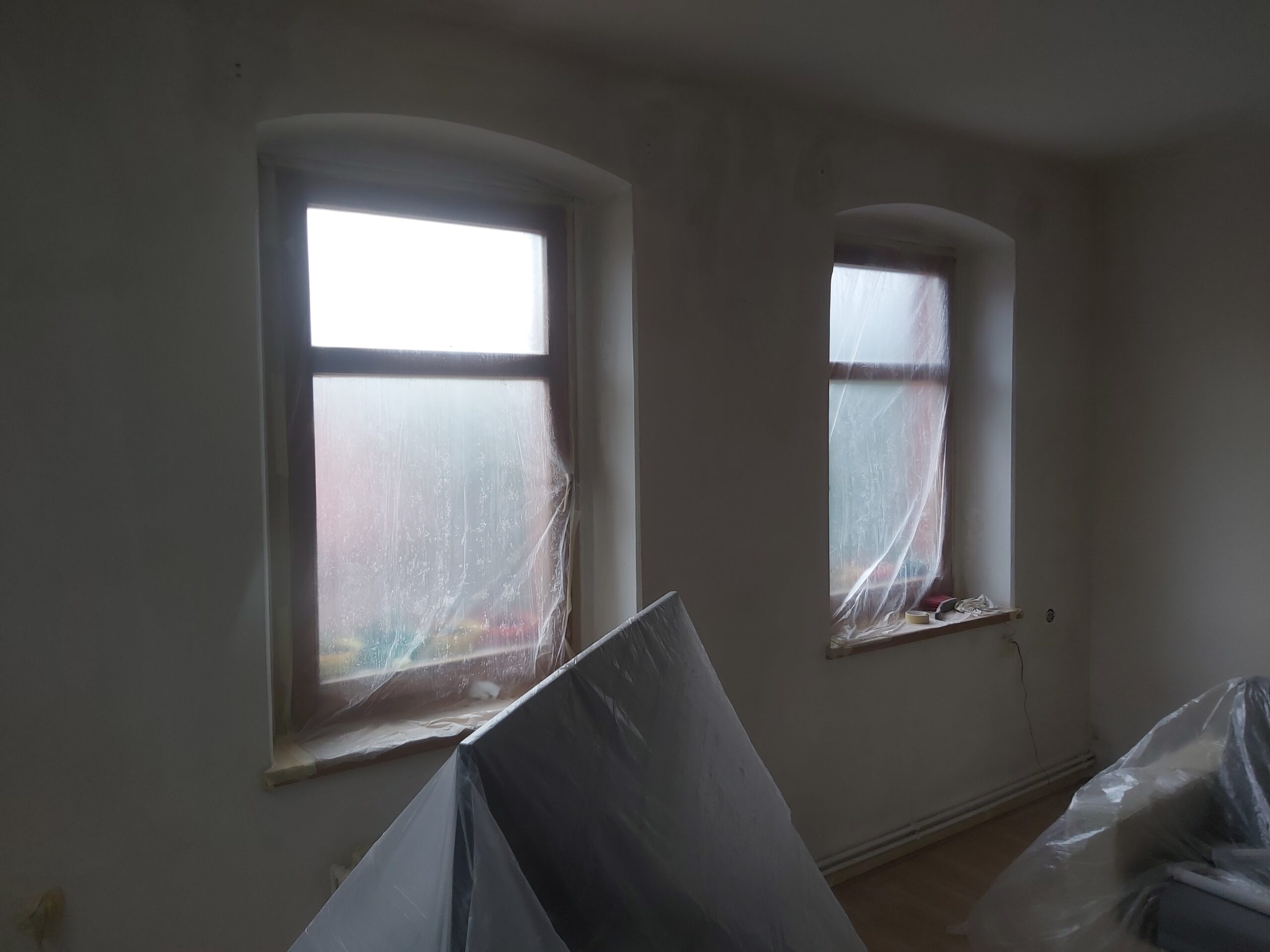 Pokój w trakcie remontu, z oknami zabezpieczonymi folią i meblami przykrytymi foliami ochronnymi.