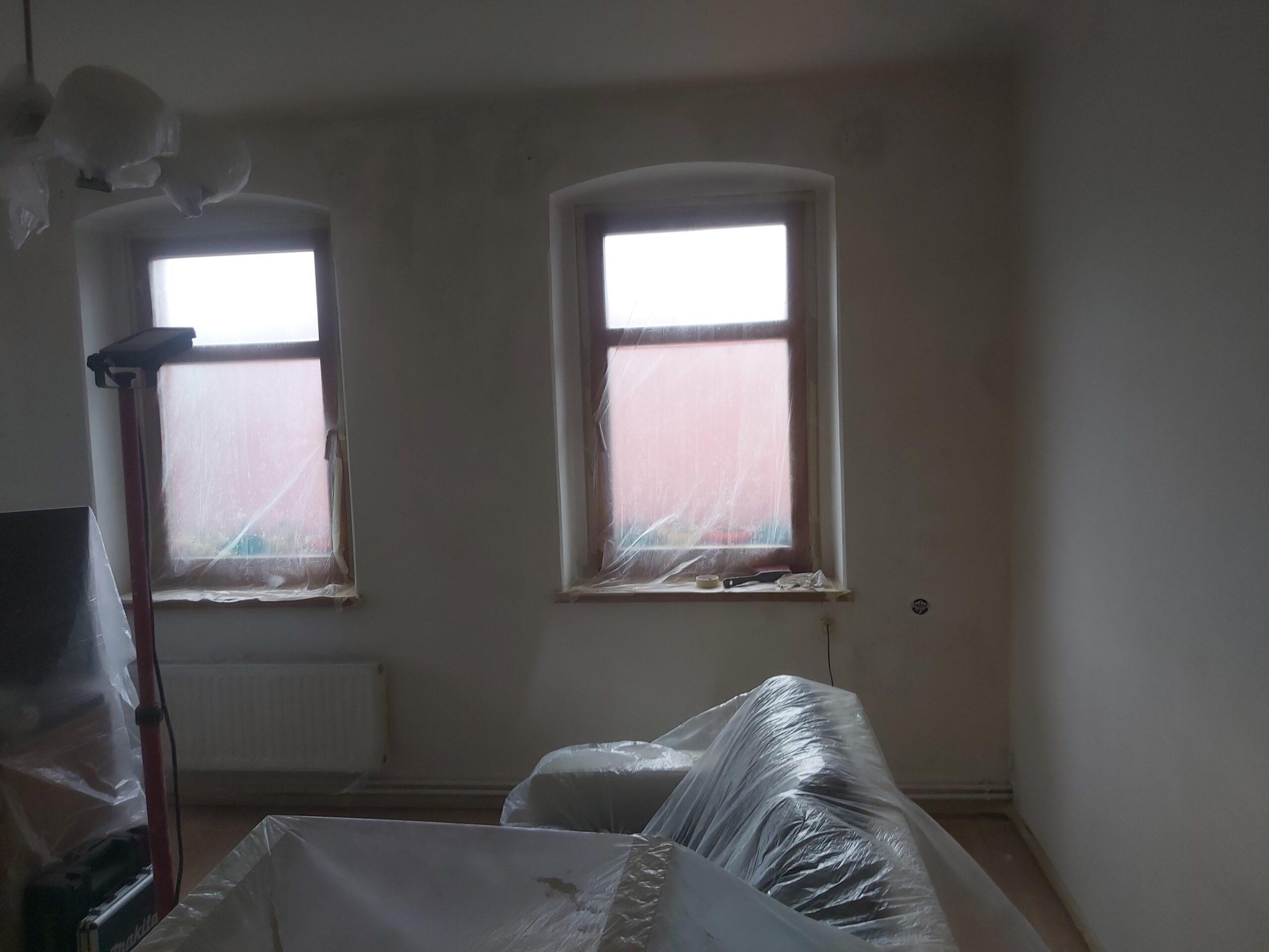 Pokój w trakcie przygotowań do malowania, z zabezpieczonymi folią meblami oraz oknami.