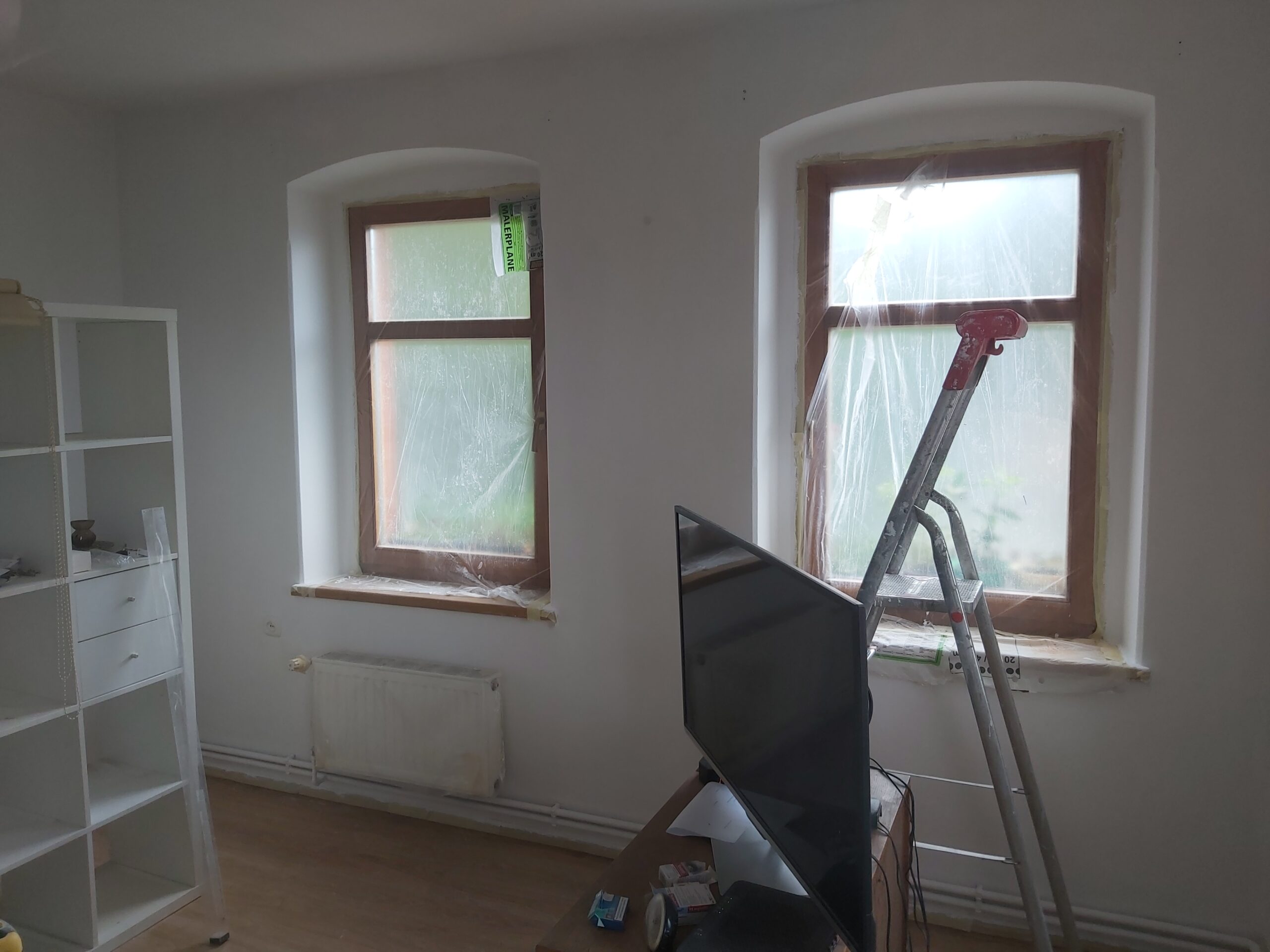 Dwa okna w pomieszczeniu podczas remontu, zabezpieczone folią, z widoczną drabiną przy jednym z nich oraz elementami wyposażenia wnętrza odsuniętymi od ścian.