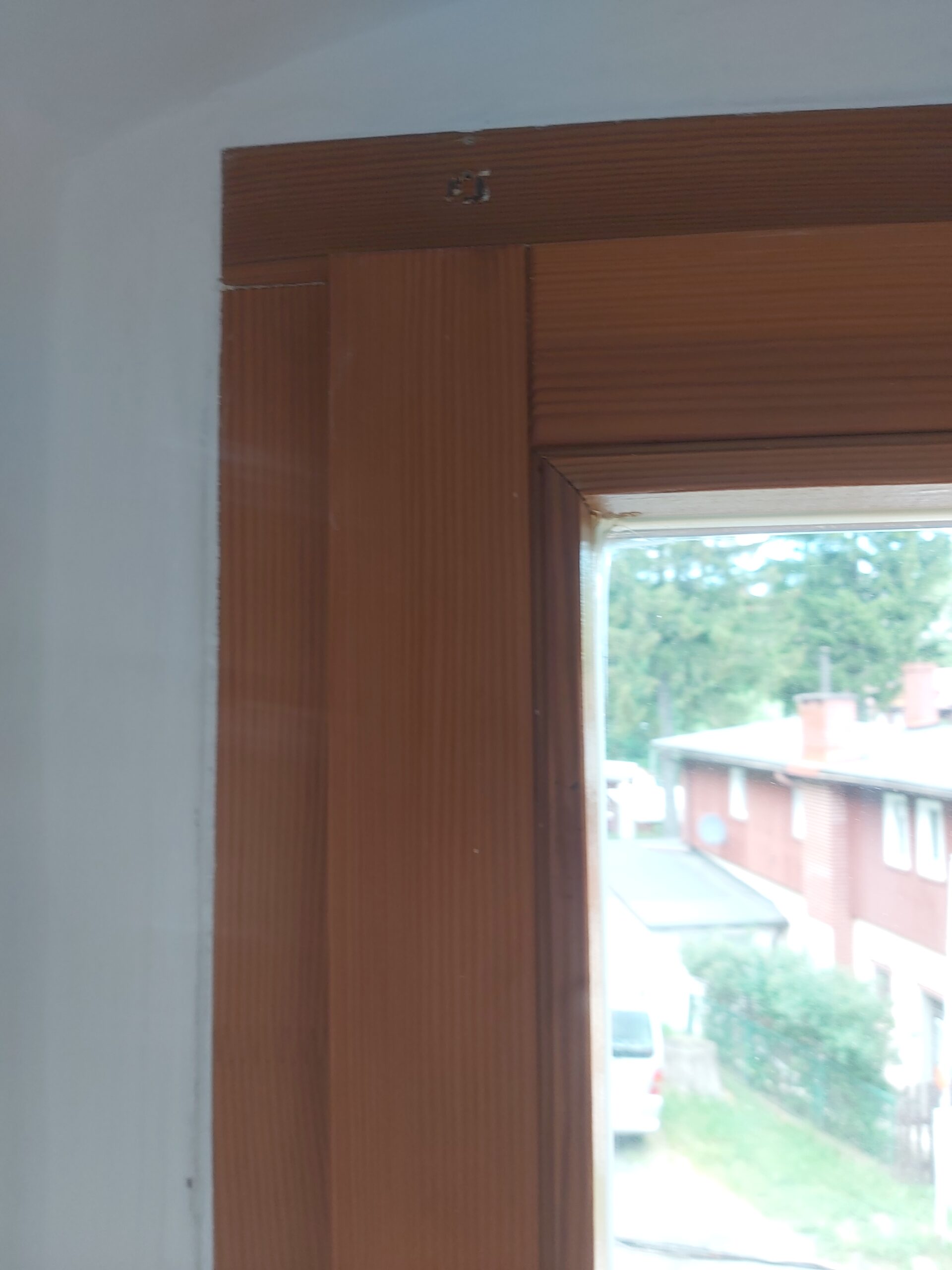 Drewniana ościeżnica okienna widziana z bliska, narożnik górny z widocznym łączeniem elementów i białą, odnowioną ścianą wokół okna.