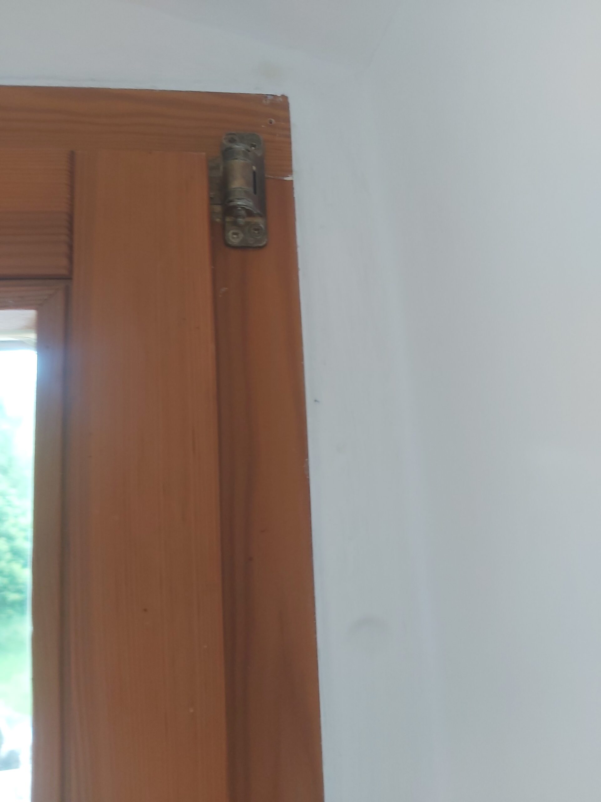 Zbliżenie na górny narożnik drewnianej ościeżnicy okiennej z widocznym zawiasem oraz świeżo pomalowaną białą ścianą po pracach wykończeniowych.