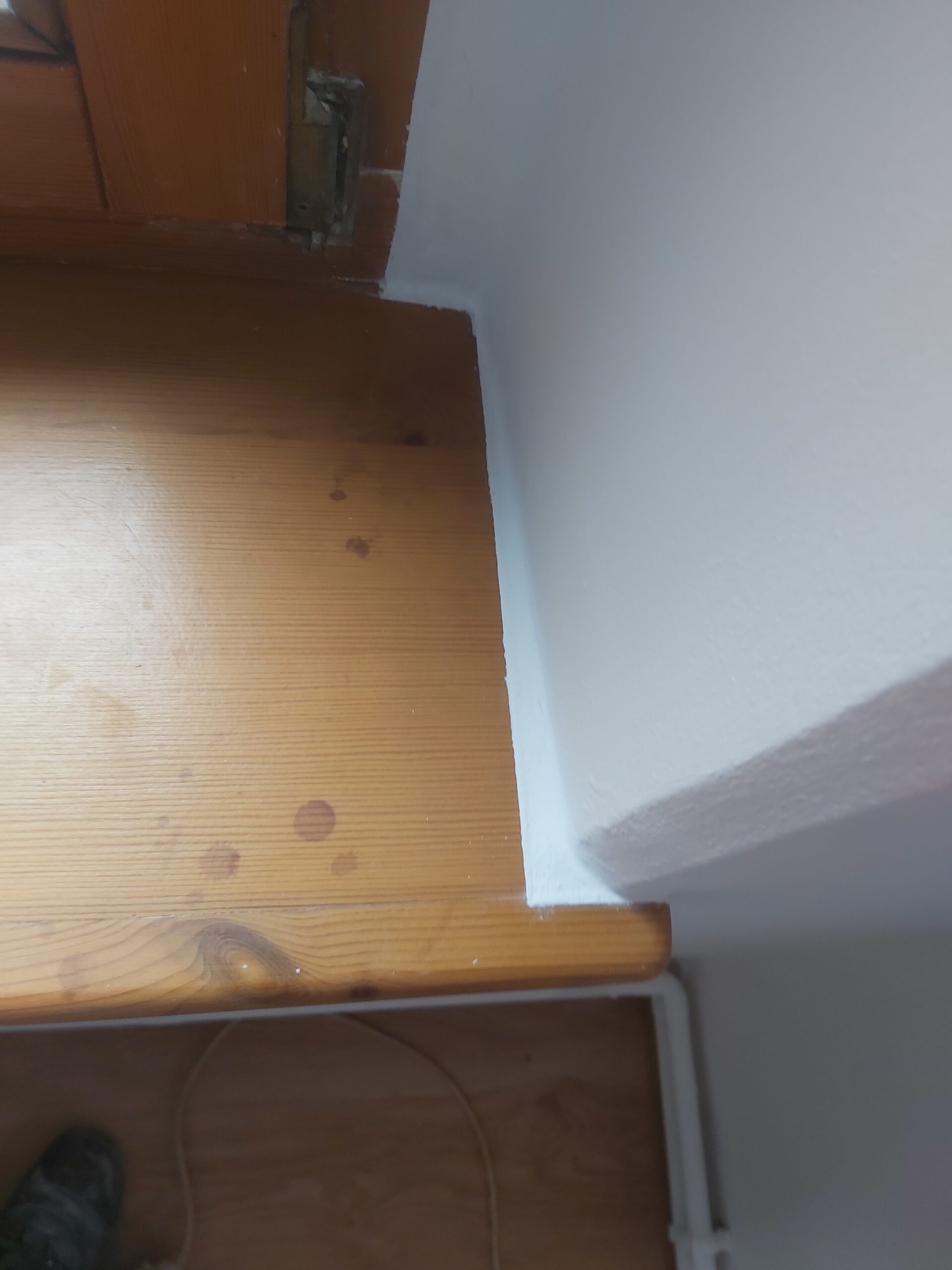 Zbliżenie na narożnik ściany i drewniany parapet po malowaniu, widoczne świeże białe wykończenie krawędzi oraz ślady po pracach remontowych.
