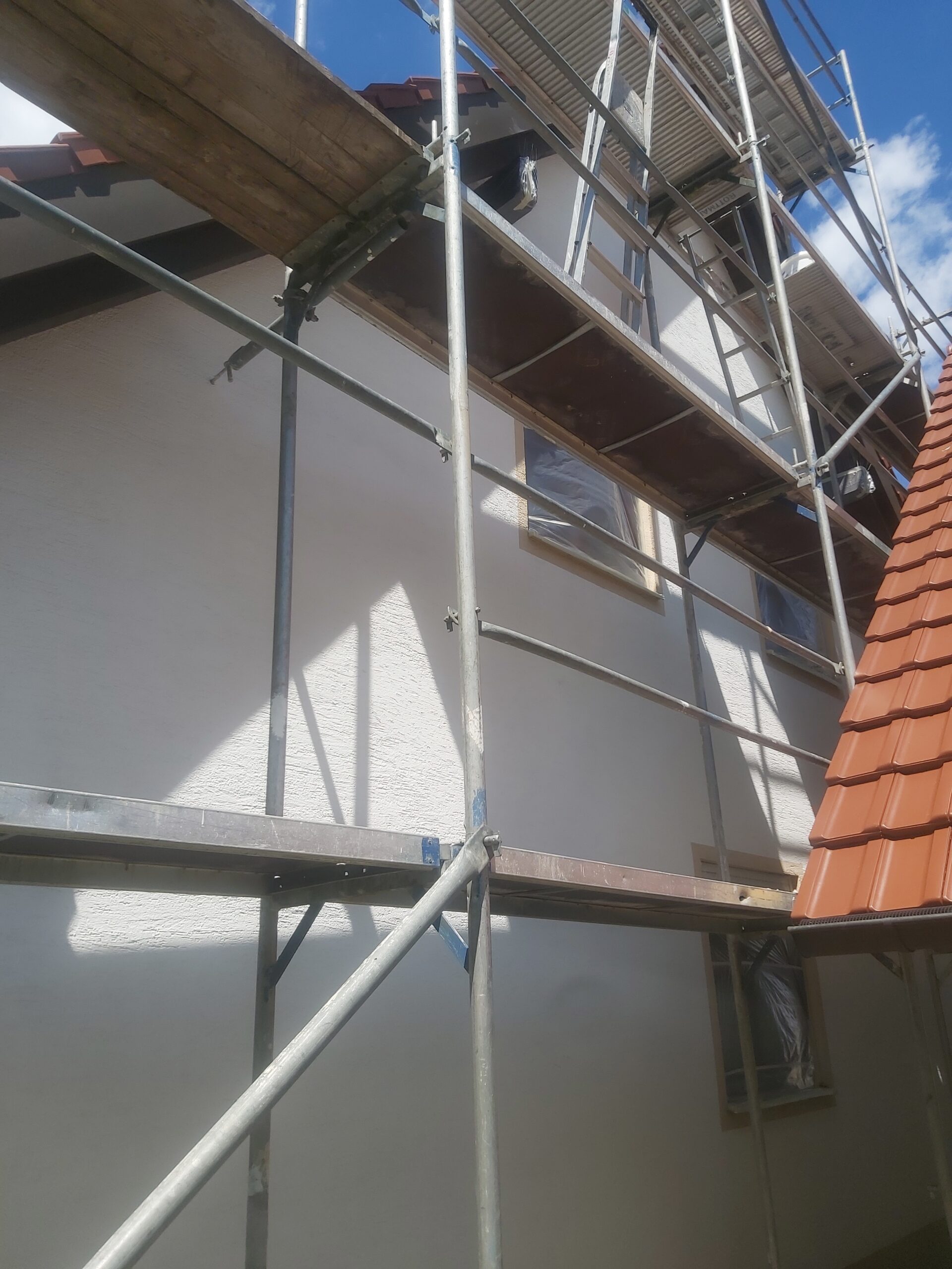 Metalowe rusztowanie ustawione przy bocznej elewacji domu, z widocznymi pomostami roboczymi i zabezpieczonym oknem podczas prowadzonych prac elewacyjnych.