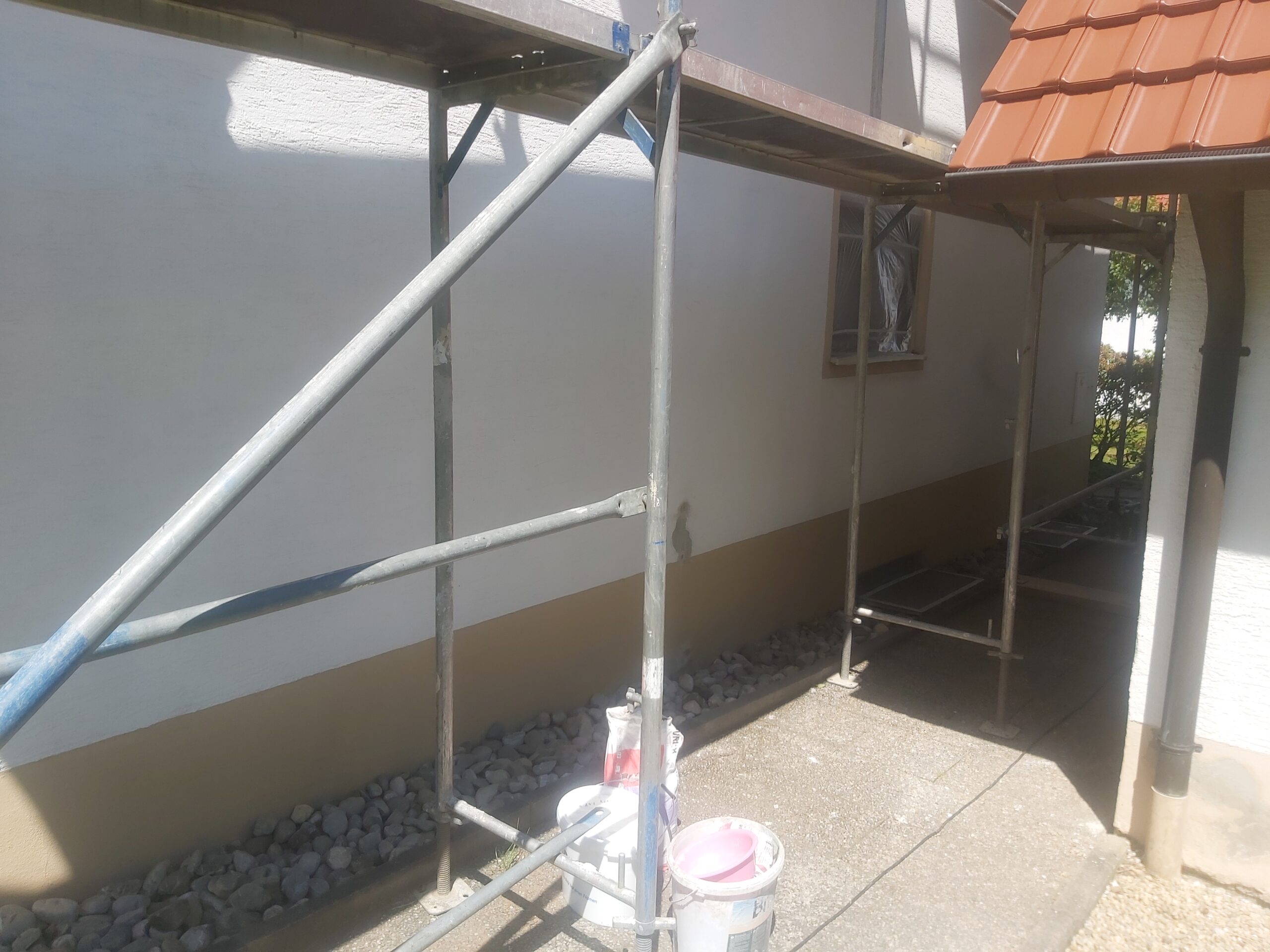 Rusztowanie ustawione wzdłuż bocznej ściany domu jednorodzinnego, z widocznymi wiadrami po farbie na podłożu i zabezpieczoną elewacją przygotowaną do prac remontowych