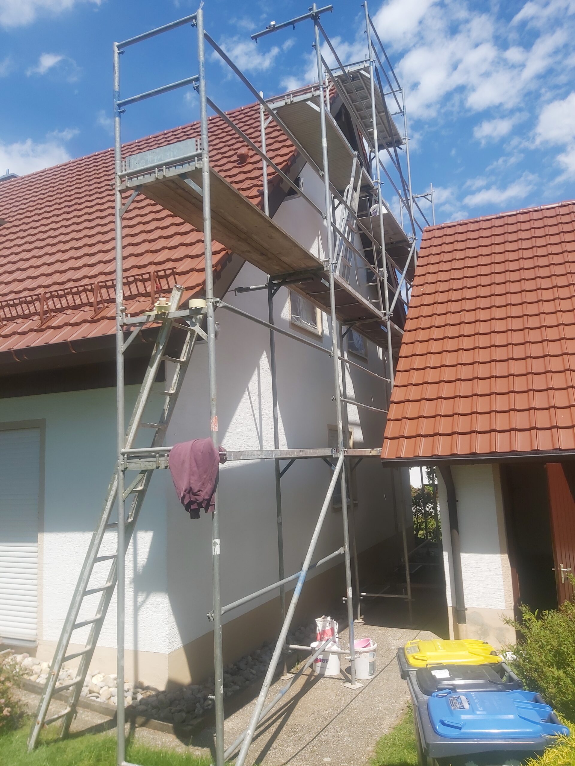 Metalowe rusztowanie ustawione przy elewacji domu jednorodzinnego, z pomostami roboczymi i drabiną, przygotowane do prac na ścianach i pod dachem.