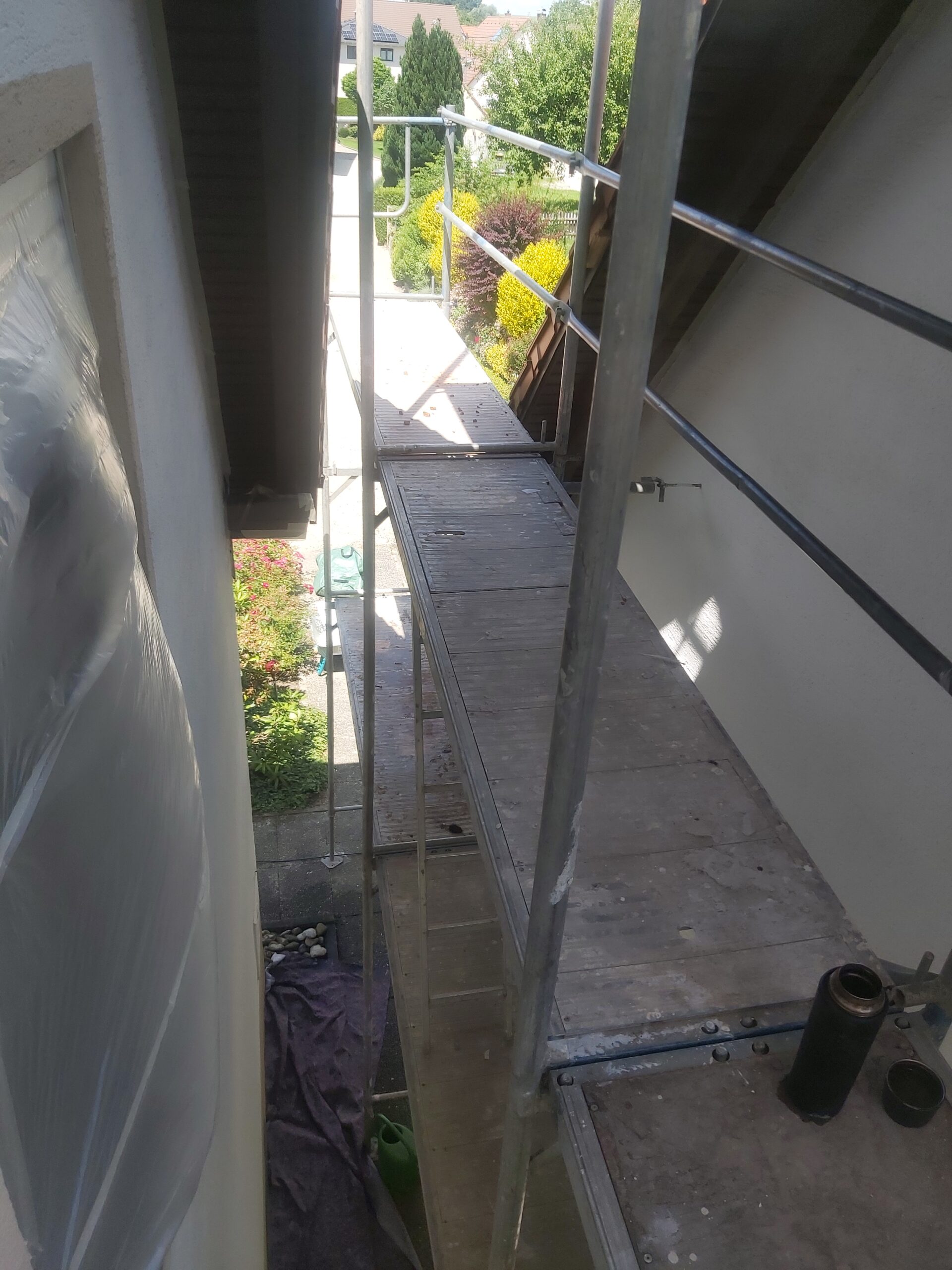 Wąski pomost roboczy metalowego rusztowania ustawionego między elewacją budynku a dachem, widok z góry, poniżej ogród i ścieżka wokół domu.