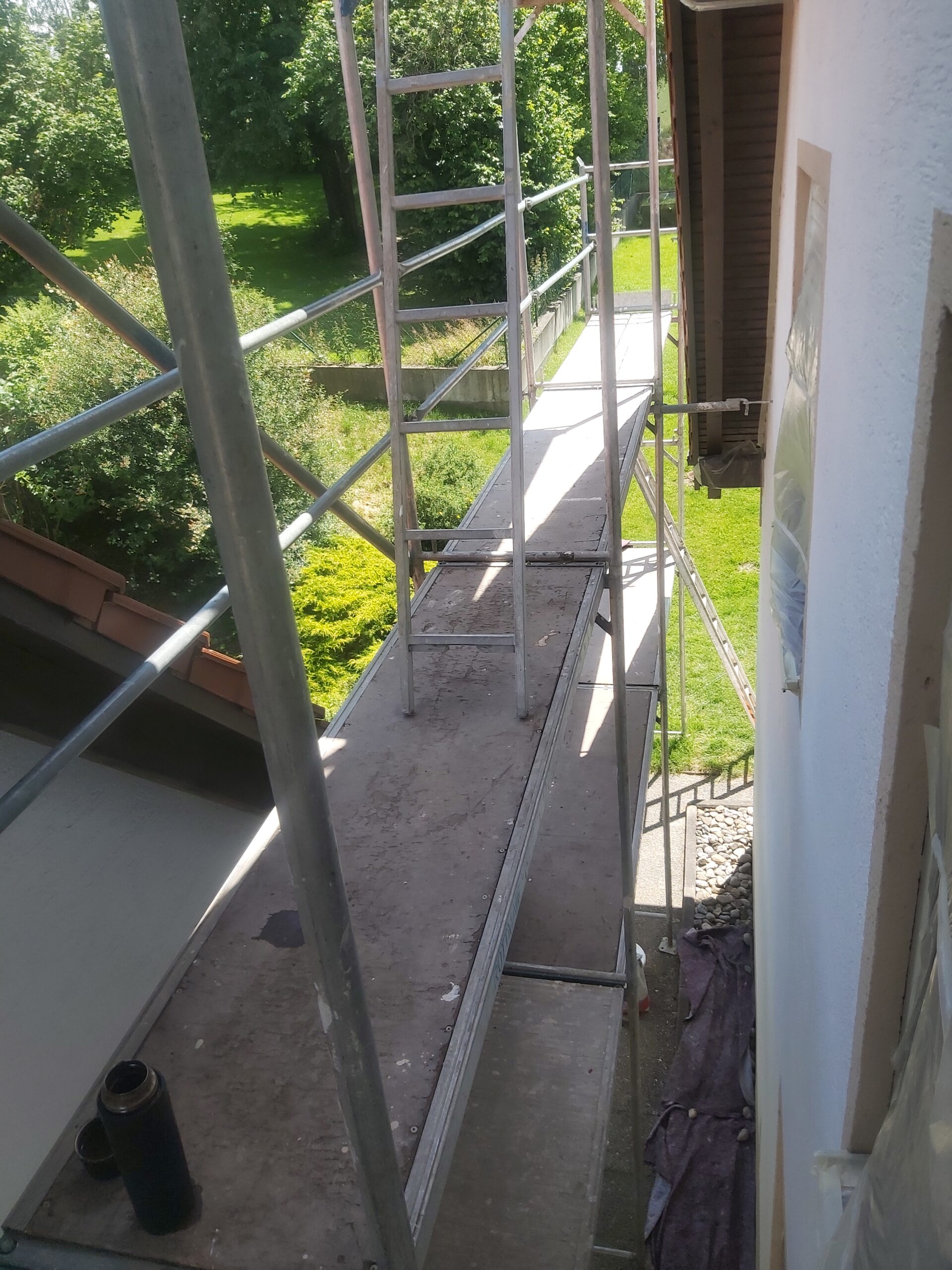 Metalowe rusztowanie ustawione wzdłuż elewacji bocznej domu, widok z poziomu pomostu roboczego, w tle zieleń ogrodu i teren wokół budynku.