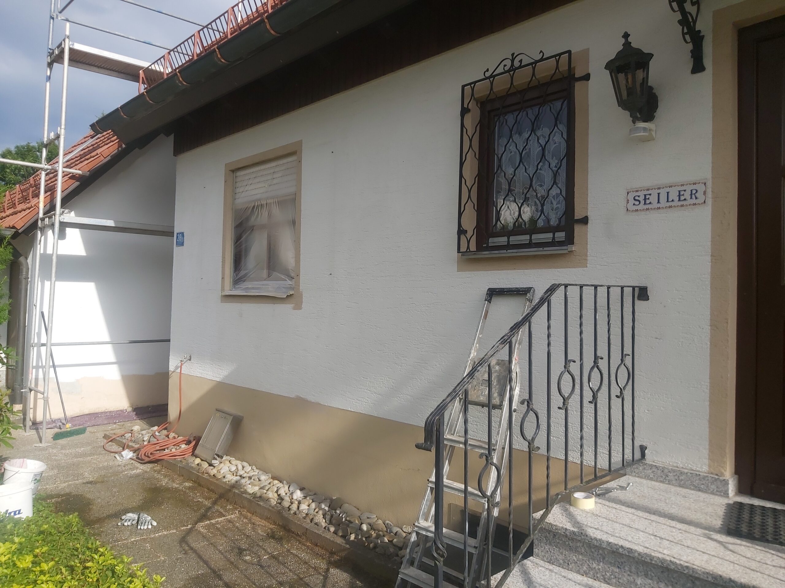 Frontowa elewacja domu jednorodzinnego w trakcie końcowych prac wykończeniowych, widoczne schody z metalową balustradą, zabezpieczone okno, lampa ścienna oraz odnowiony cokół elewacji.