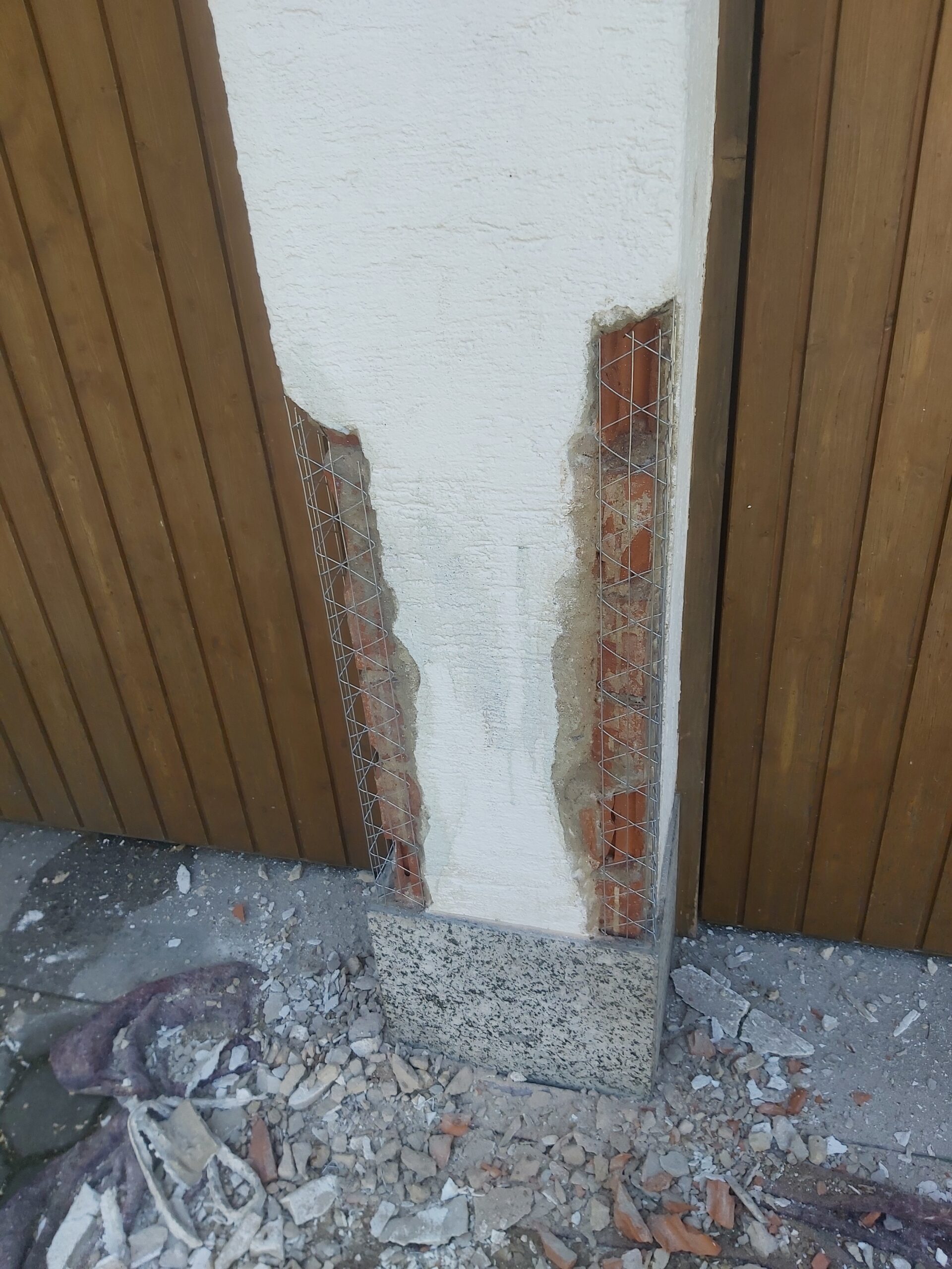 Słup elewacyjny między drewnianymi bramami garażowymi w trakcie naprawy, odsłonięta cegła zabezpieczona siatką zbrojącą oraz świeżo nałożona warstwa tynku.