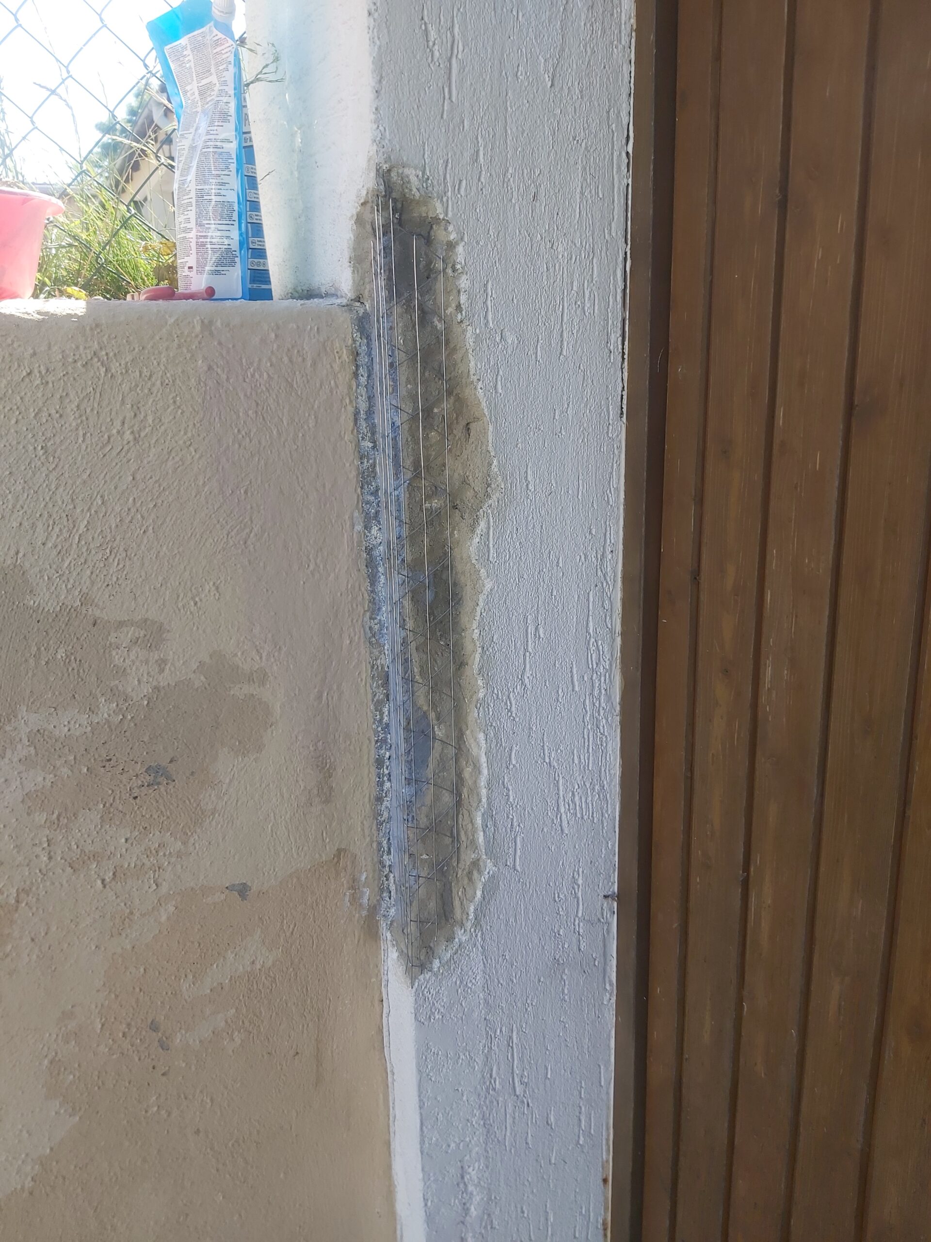 Zbliżenie na narożnik zewnętrznej ściany budynku przy drewnianej bramie garażowej, z widoczną siatką zbrojącą i świeżo nałożoną zaprawą podczas prac naprawczych elewacji.