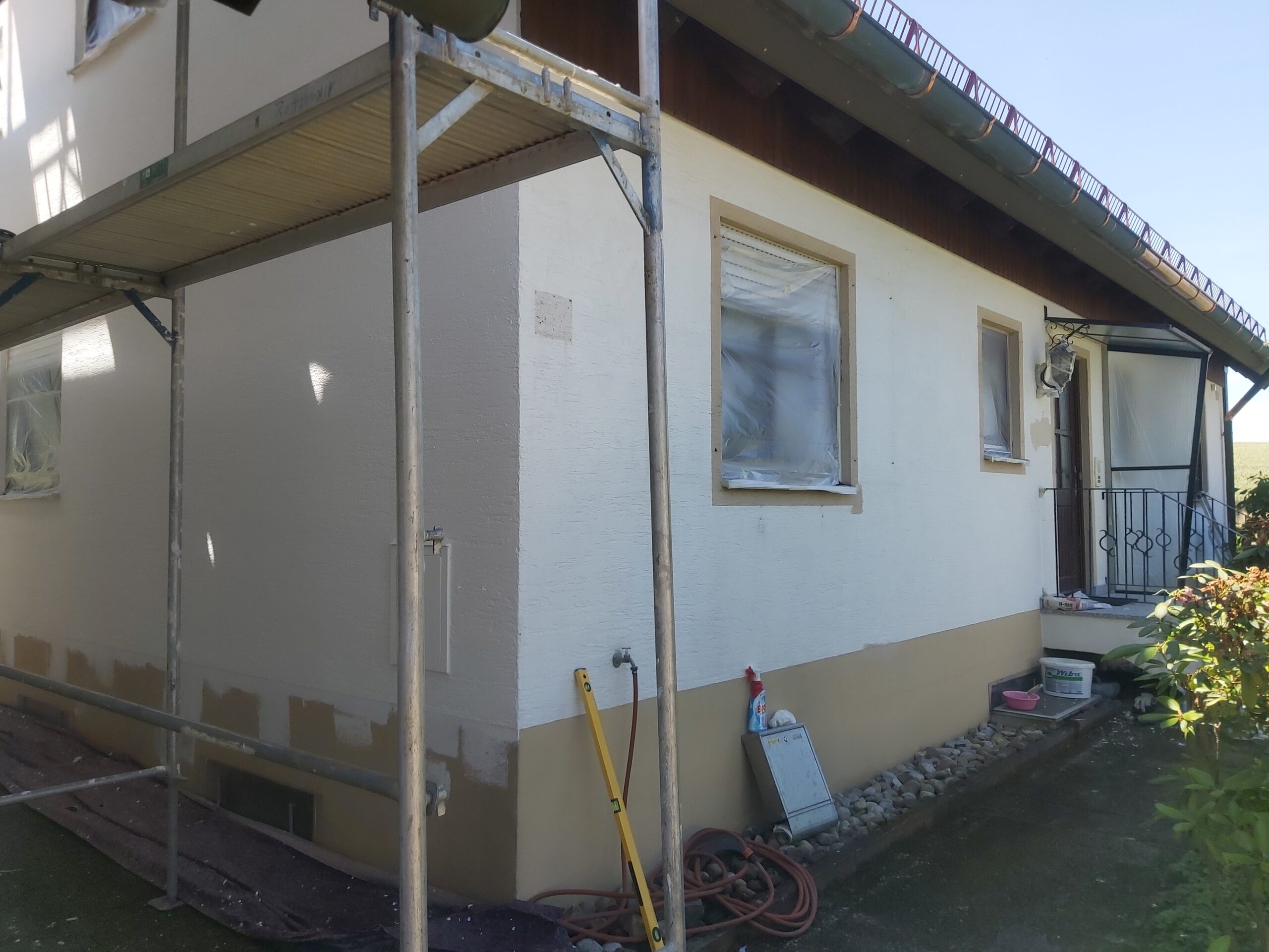 Boczne ujęcie elewacji domu jednorodzinnego podczas remontu, widoczne rusztowanie, zabezpieczone okna folią, odnowiony cokół oraz narzędzia budowlane przy ścianie.