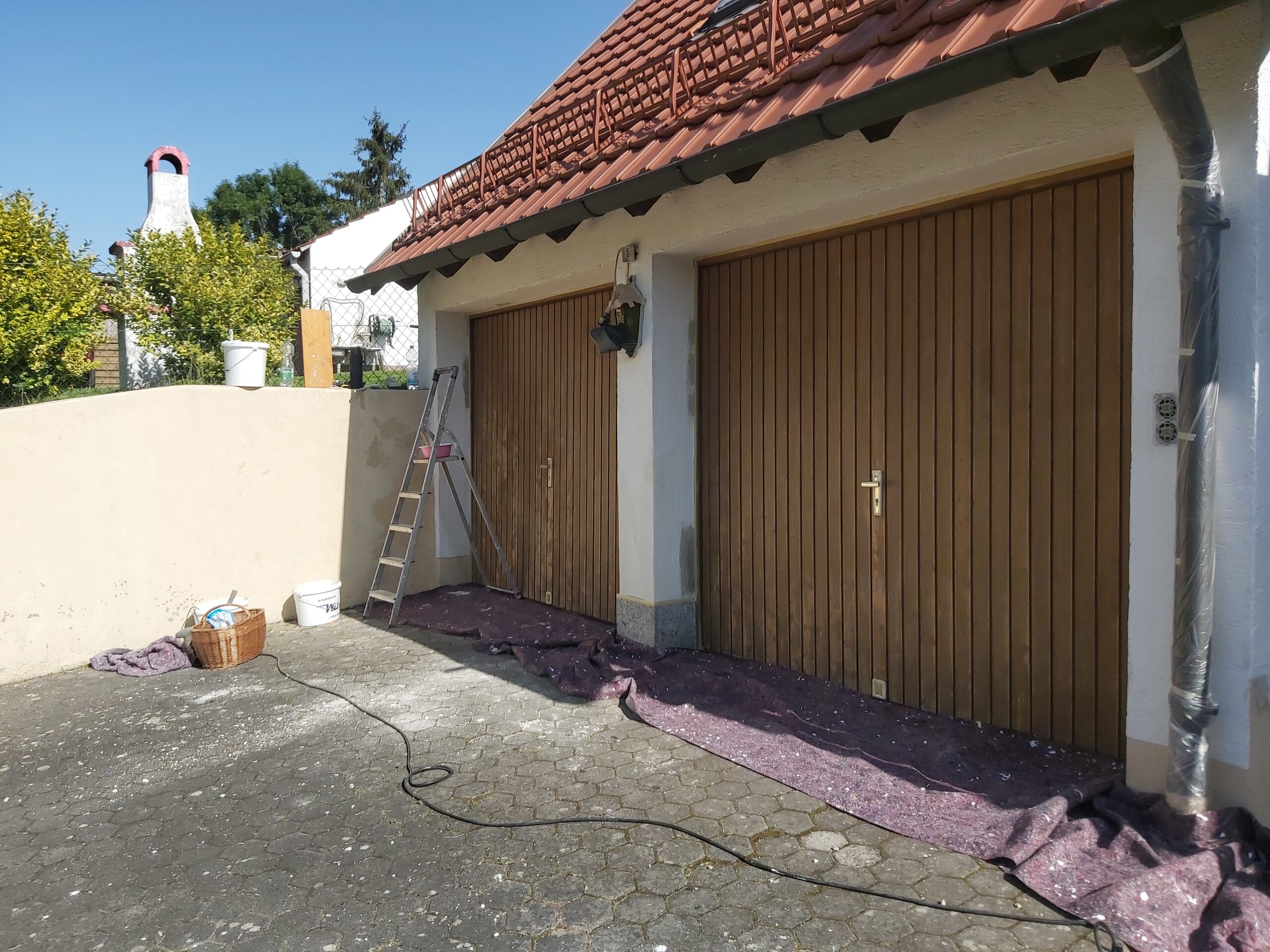 Elewacja budynku z dwoma drewnianymi drzwiami garażowymi w trakcie malowania, widoczna drabina, zabezpieczone podłoże oraz przygotowanie do prac wykończeniowych.