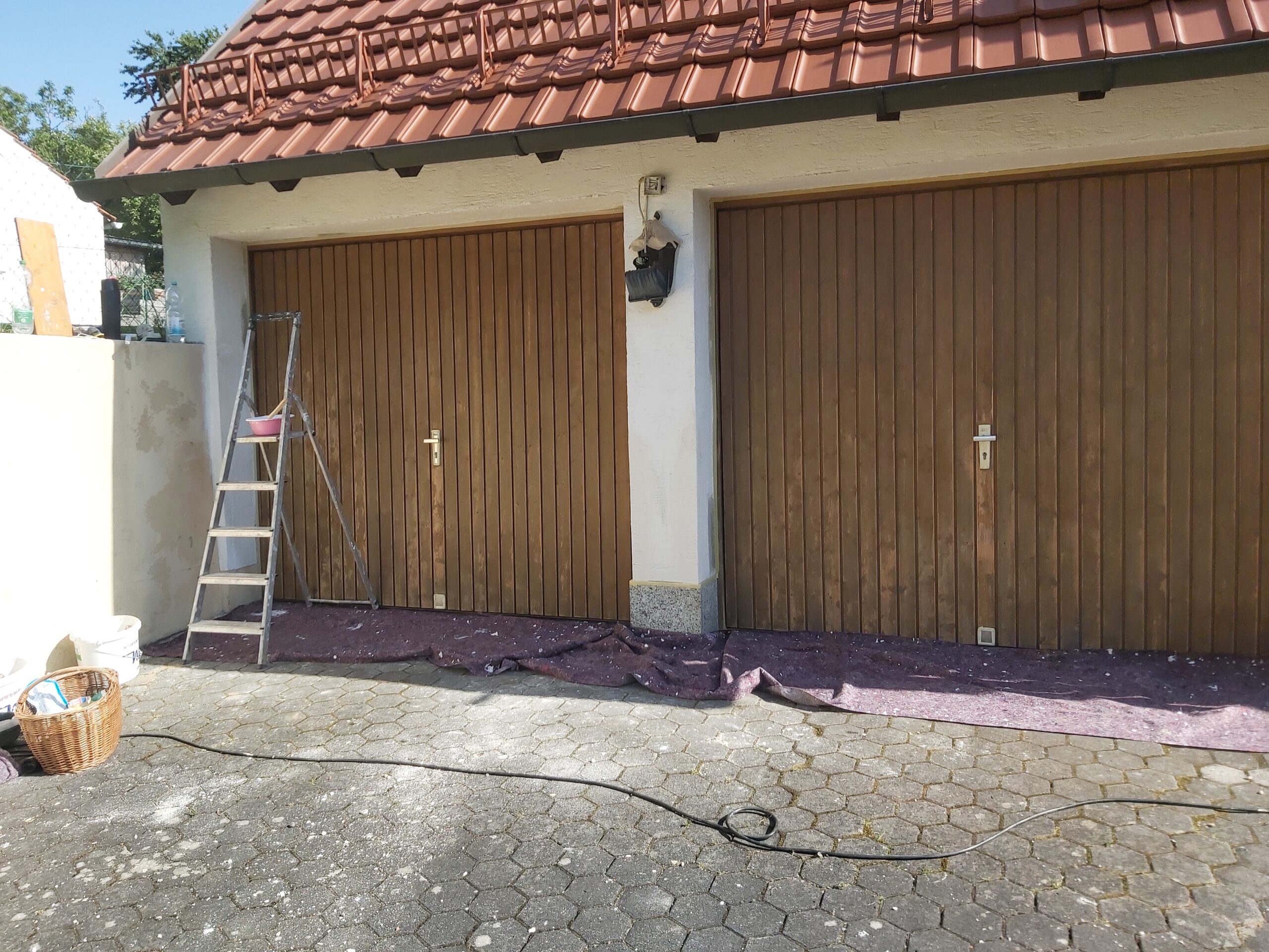 Dwie drewniane bramy garażowe przed domem, zabezpieczone od dołu folią ochronną, z drabiną ustawioną przy lewej bramie i narzędziami przygotowanymi do prac renowacyjnych.