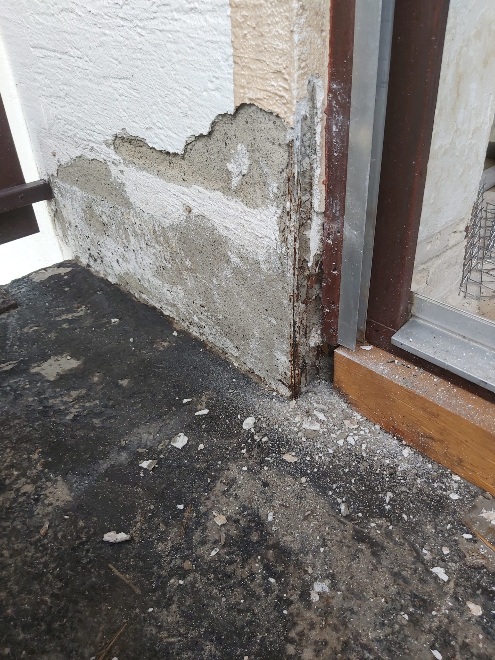 Uszkodzony i kruszący się tynk elewacyjny w narożniku przy progu drzwi balkonowych, widoczne zawilgocenie, odsłonięta warstwa podkładowa oraz zabrudzenia przy cokole.