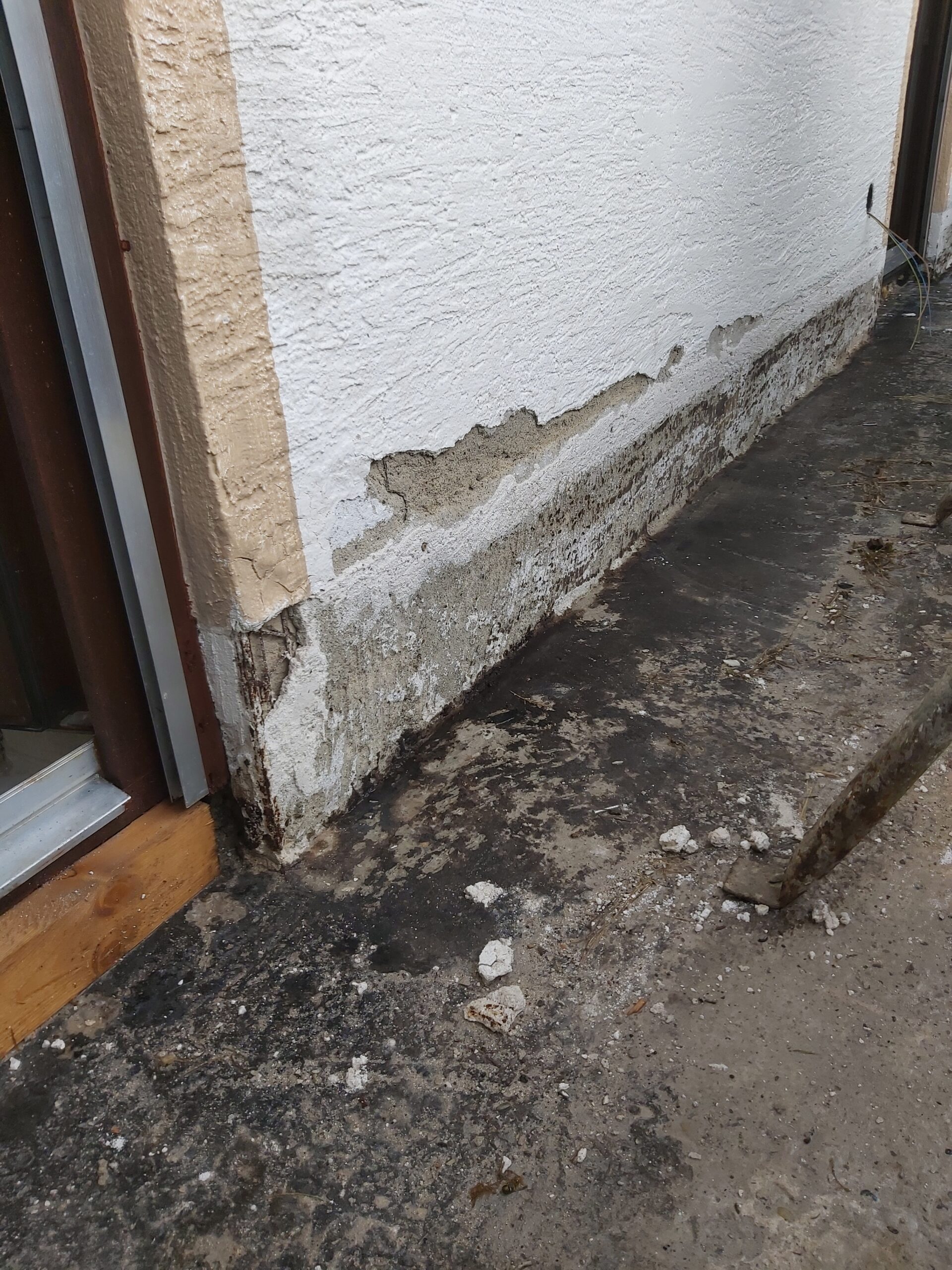 Odpadający tynk i zawilgocony fragment elewacji przy cokole budynku, widoczne ubytki oraz odsłonięta warstwa podkładowa przy wejściu.