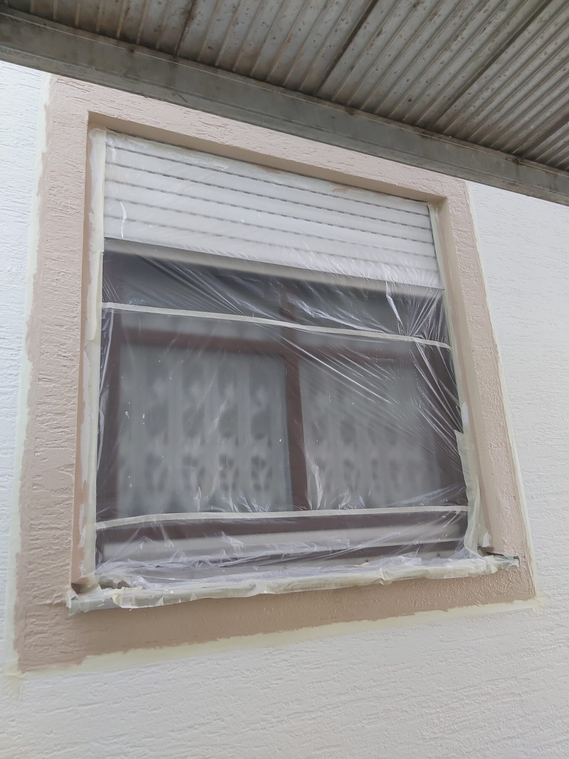 Okno z roletą zewnętrzną zabezpieczone folią i taśmą malarską, widoczna świeżo malowana opaska wokół okna na elewacji domu.