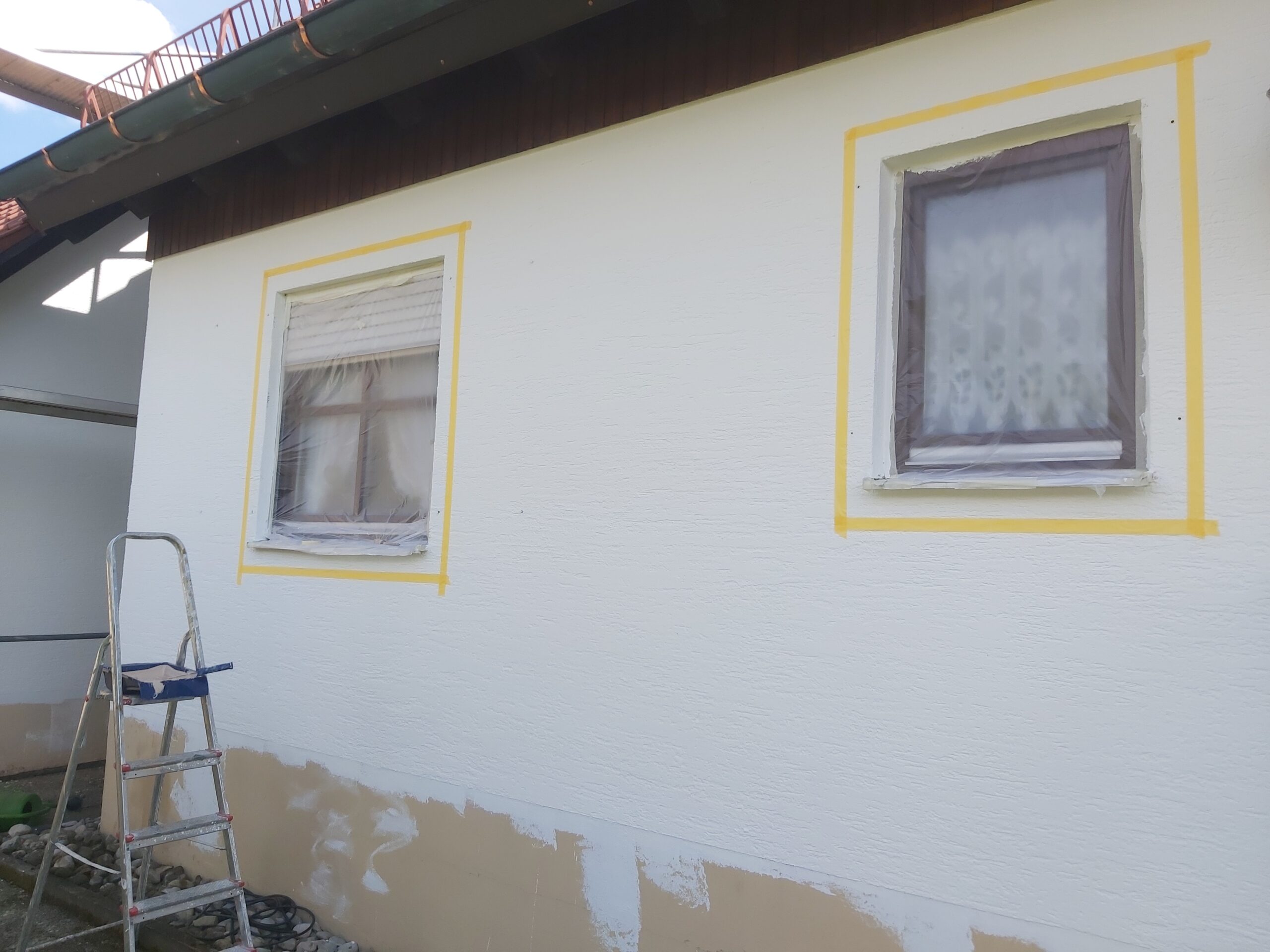 Fragment elewacji domu z dwoma oknami zabezpieczonymi taśmą malarską, drabina ustawiona przy ścianie, widoczne ślady prac remontowych przy dolnej części elewacji.