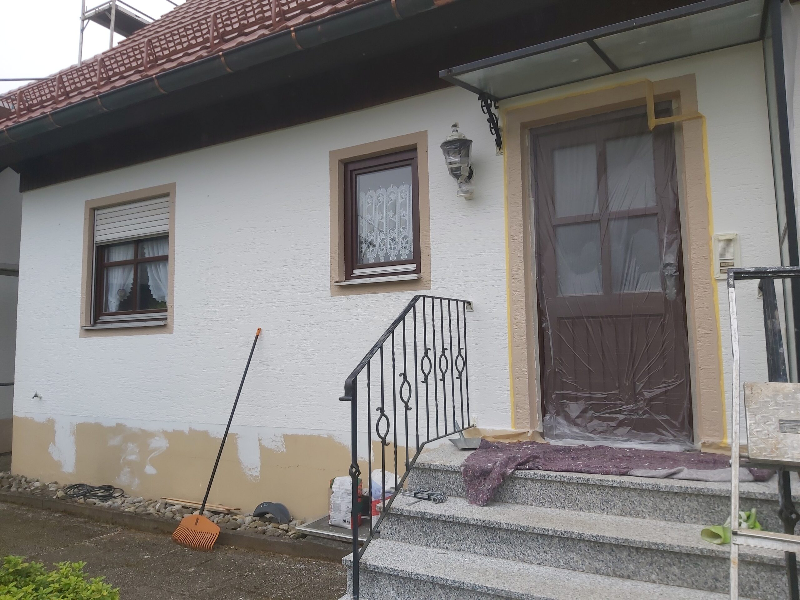 Fragment elewacji domu jednorodzinnego w trakcie remontu, widoczne wejście z drzwiami zabezpieczonymi folią, schody z balustradą oraz dolna część ściany przygotowana do dalszych prac tynkarskich.