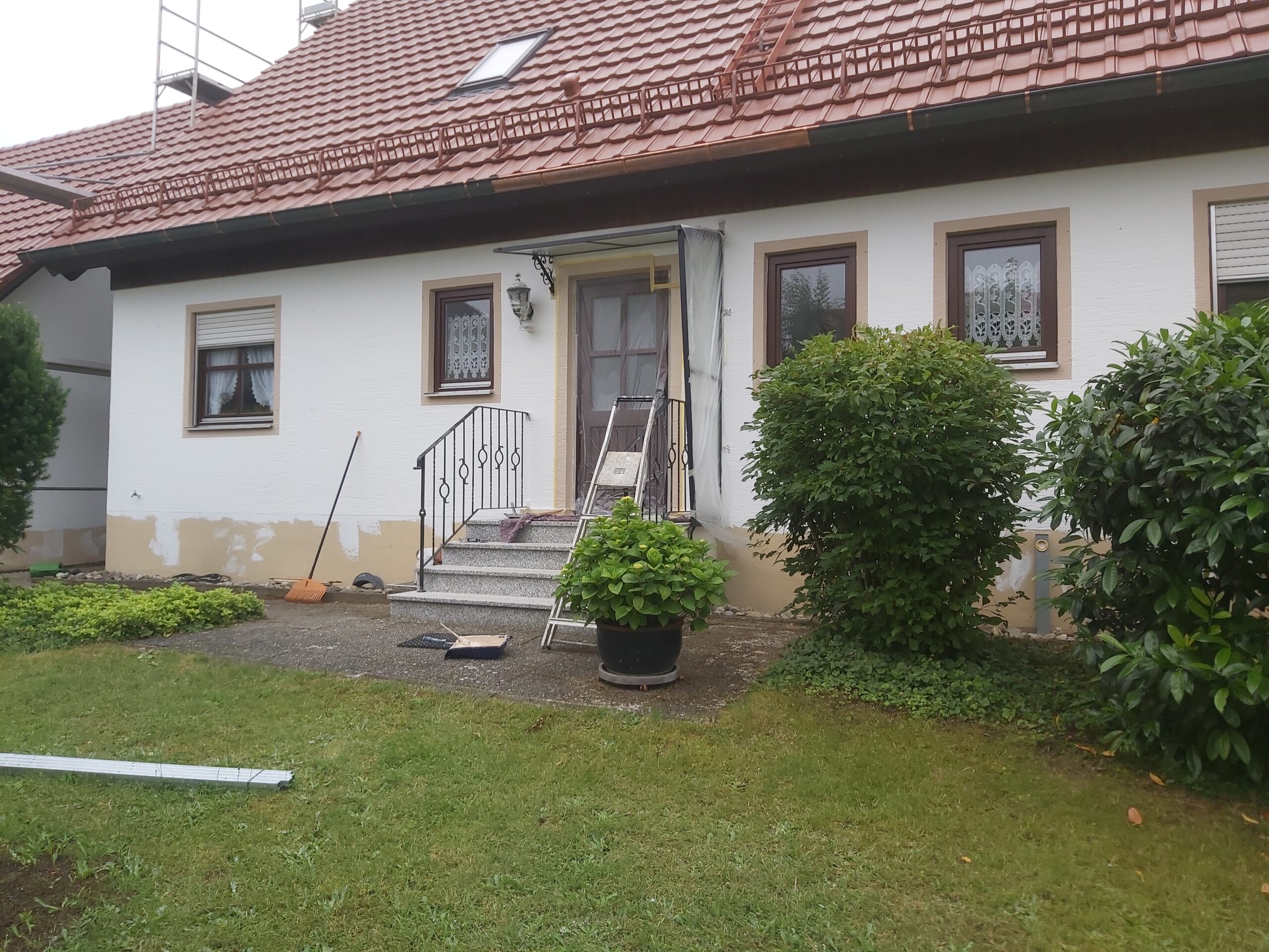 Frontowa elewacja domu jednorodzinnego podczas prac remontowych, widoczne zabezpieczenia, drabina przy wejściu oraz fragmenty naprawianego tynku u podstawy ściany.