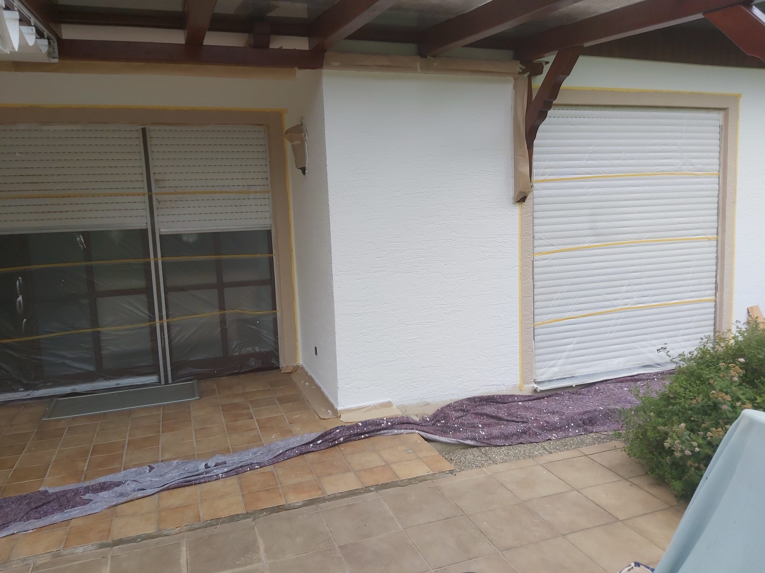 Fragment elewacji przy tarasie z zabezpieczonymi oknami i roletami zewnętrznymi, widoczna świeżo wykończona ściana oraz zabezpieczenia malarskie na posadzce.