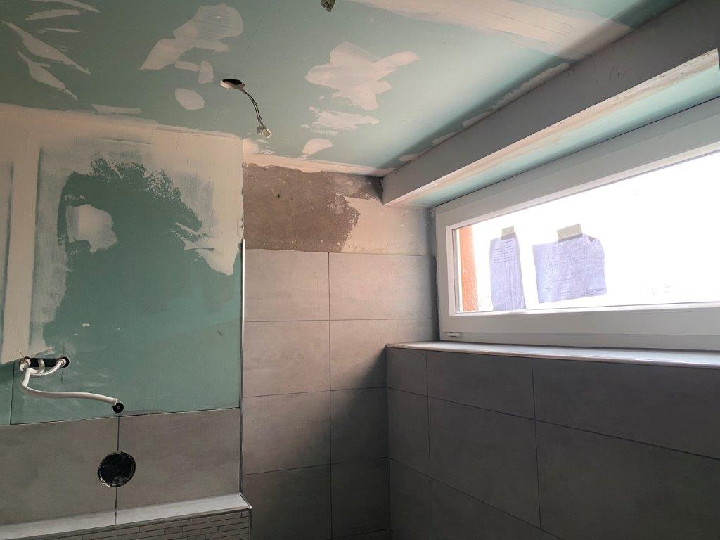 Montaż szarych płytek ściennych w łazience, widok narożnika z oknem oraz sufitu w trakcie prac wykończeniowych.