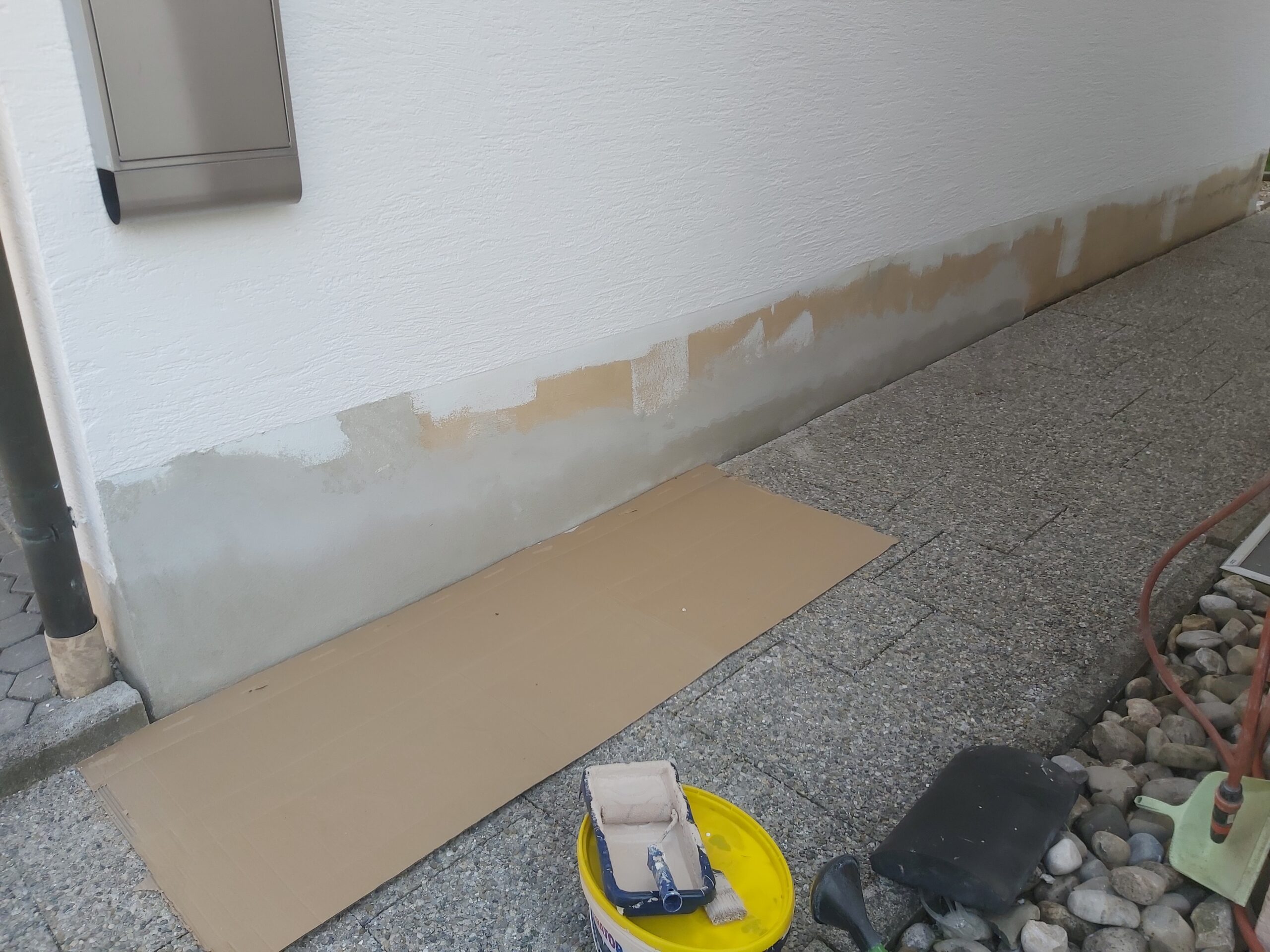 Fragment elewacji budynku z naprawianym cokołem, widoczne miejsca uzupełnień tynku oraz przygotowane podłoże zabezpieczone kartonem przy chodniku.