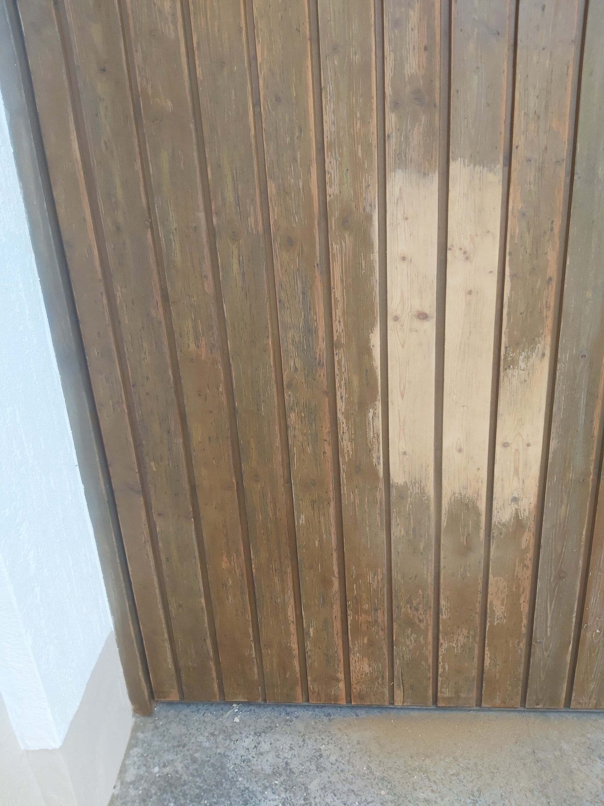 Zbliżenie na drewnianą bramę garażową z widocznym jaśniejszym pasem po czyszczeniu na tle ciemniejszego, nieodnowionego drewna.