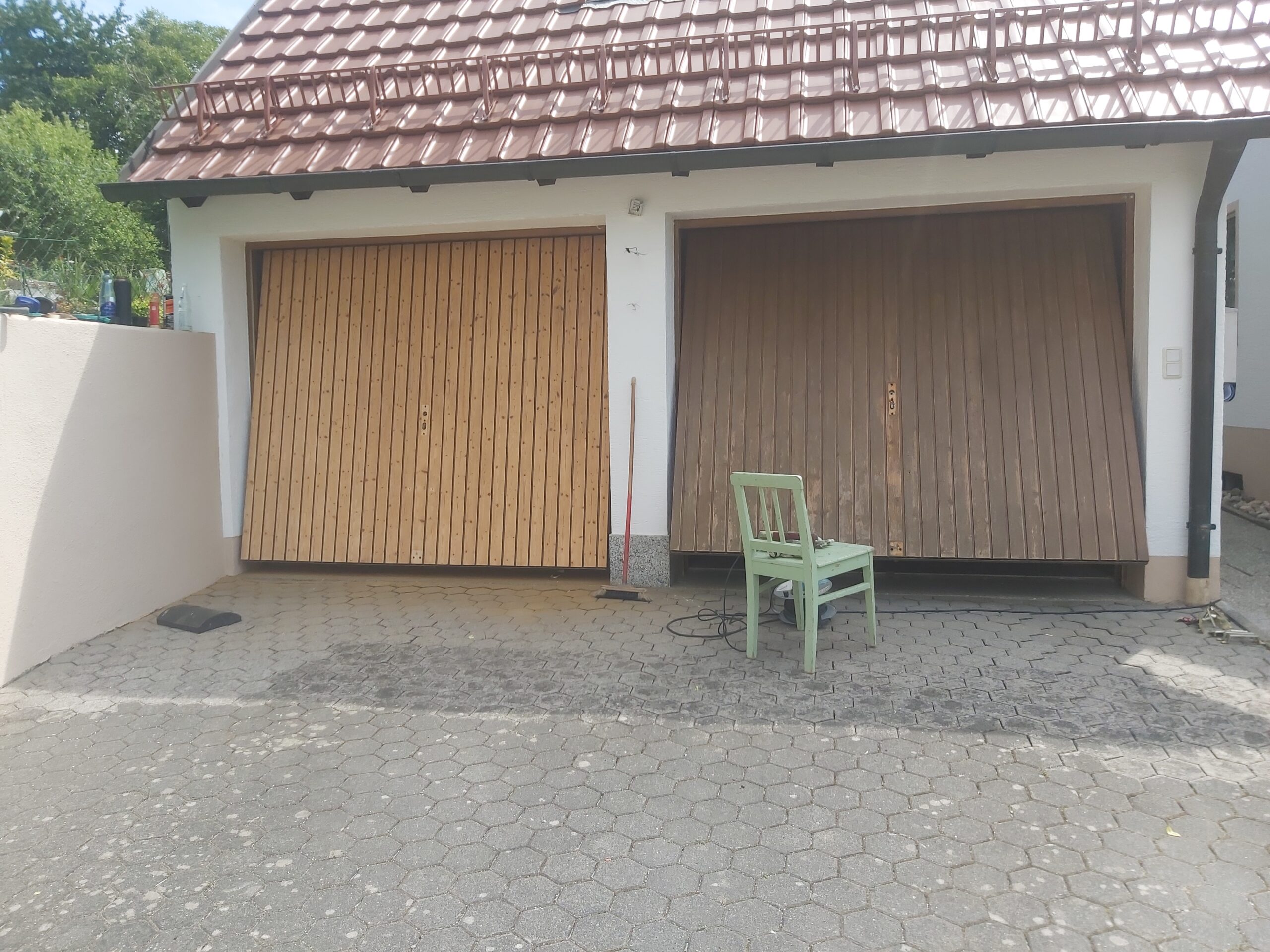 Dwie drewniane bramy garażowe obok siebie, jedna jaśniejsza po czyszczeniu, druga ciemniejsza przed odnowieniem, widok na podjazd z kostki brukowej.