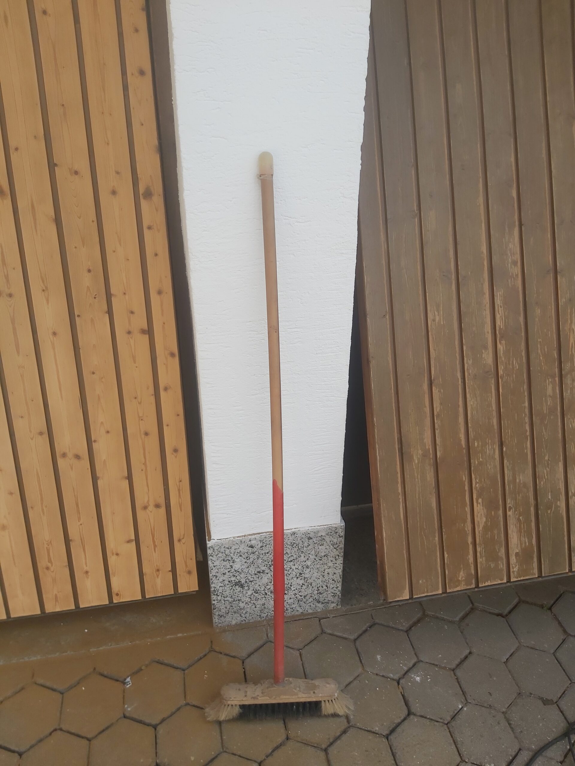 Fragment dwóch drewnianych bram garażowych o różnym odcieniu drewna, pokazujący efekt czyszczenia lub odnawiania, między bramami oparta szczotka na podjeździe z kostki.