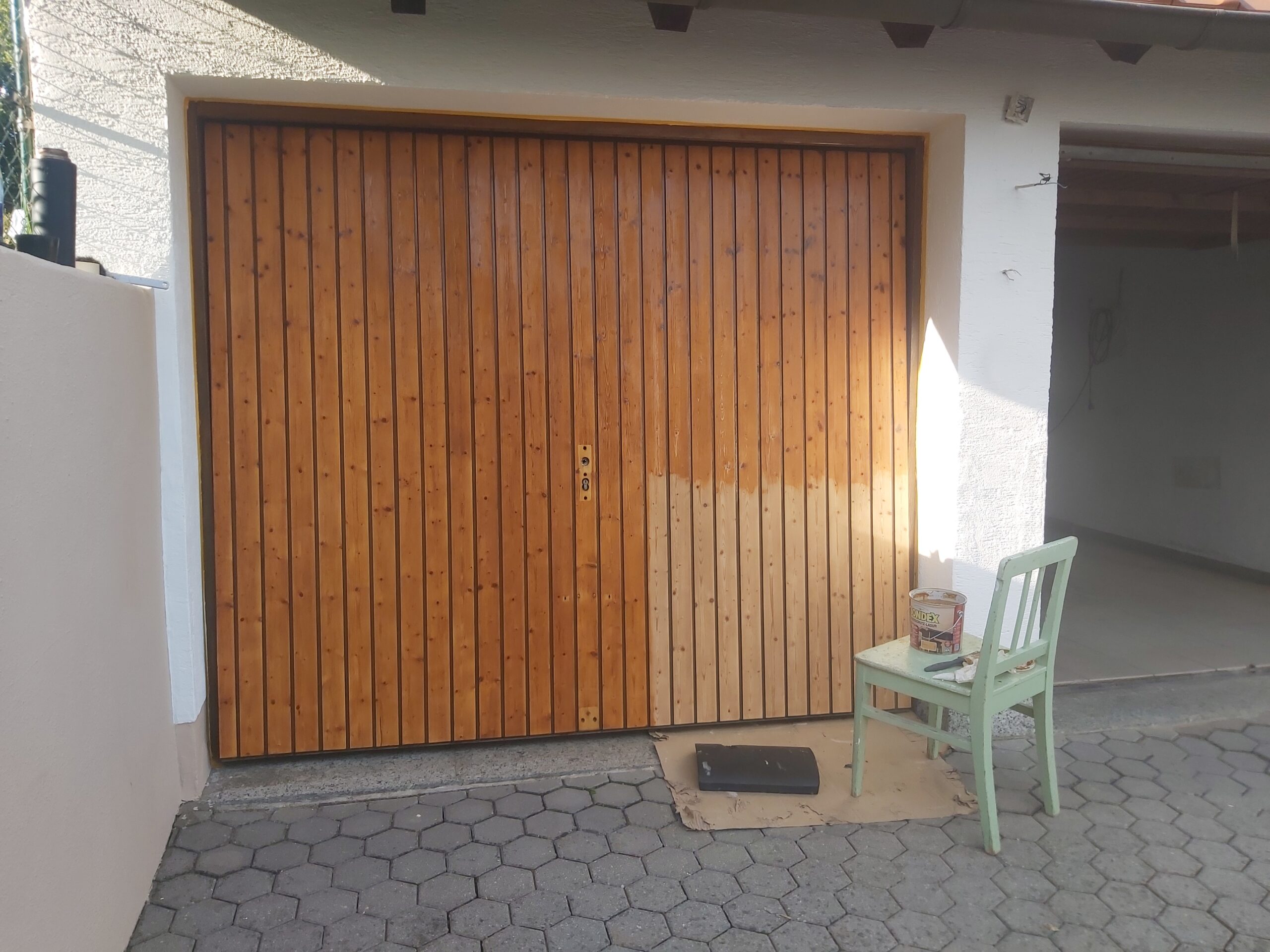 Drewniana brama garażowa z widocznym fragmentem po czyszczeniu lub szlifowaniu, zestawienie surowego i starego wykończenia drewna, przed garażem na podjeździe.