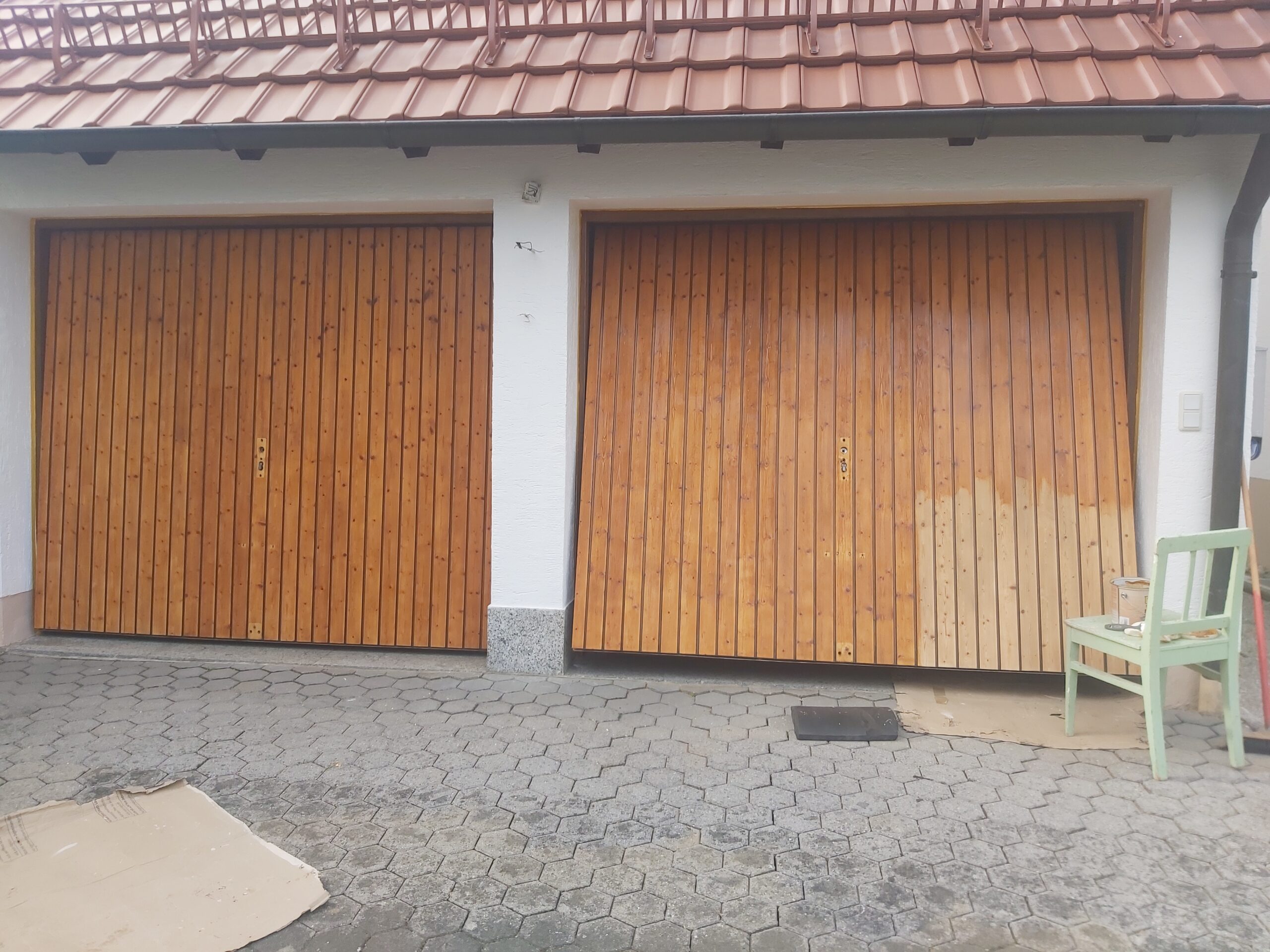 Dwie drewniane bramy garażowe przed domem, widoczne różnice w kolorze i zużyciu powierzchni drewna, ujęcie na podjeździe z kostki brukowej.