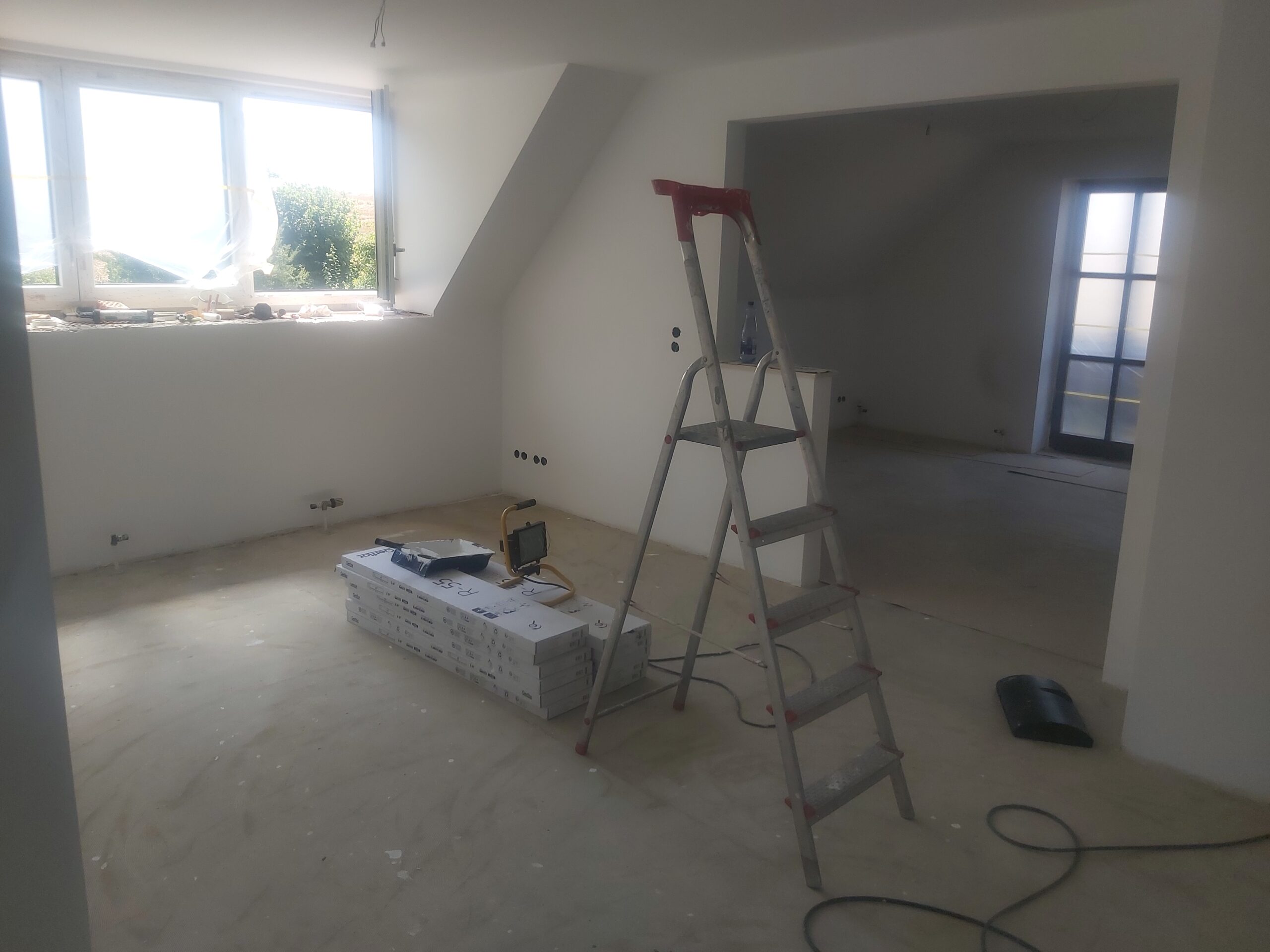 Pomieszczenie podczas remontu, z widoczną drabiną, oknem dachowym, materiałami budowlanymi na podłodze oraz otwartym przejściem do kolejnego pokoju.