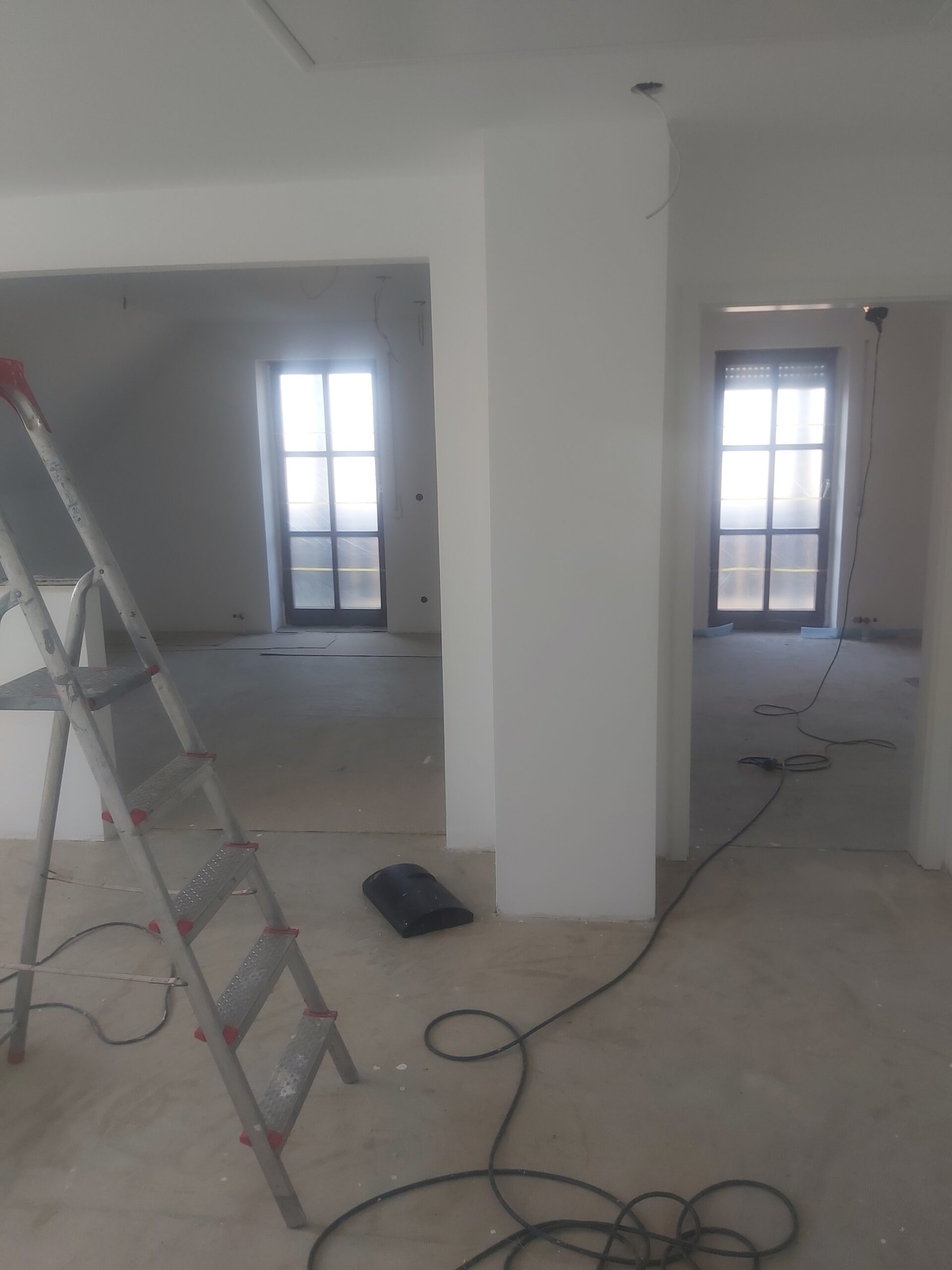 Wnętrze mieszkania w trakcie remontu, z widoczną drabiną, kolumną konstrukcyjną, przewodami elektrycznymi na podłodze oraz dwoma pomieszczeniami z drzwiami balkonowymi w tle.