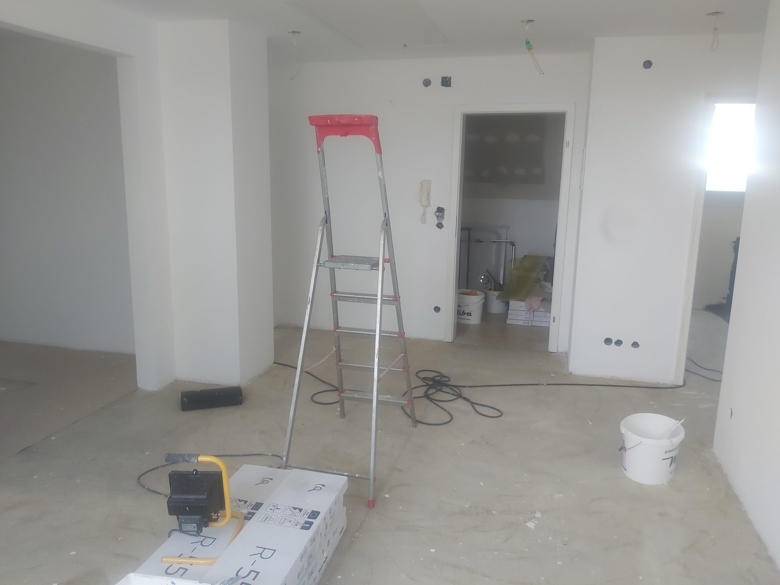 Wnętrze mieszkania w trakcie prac wykończeniowych, z białymi ścianami, drabiną stojącą na środku pomieszczenia, narzędziami, przewodami elektrycznymi oraz wiadrem i materiałami budowlanymi na podłodze.