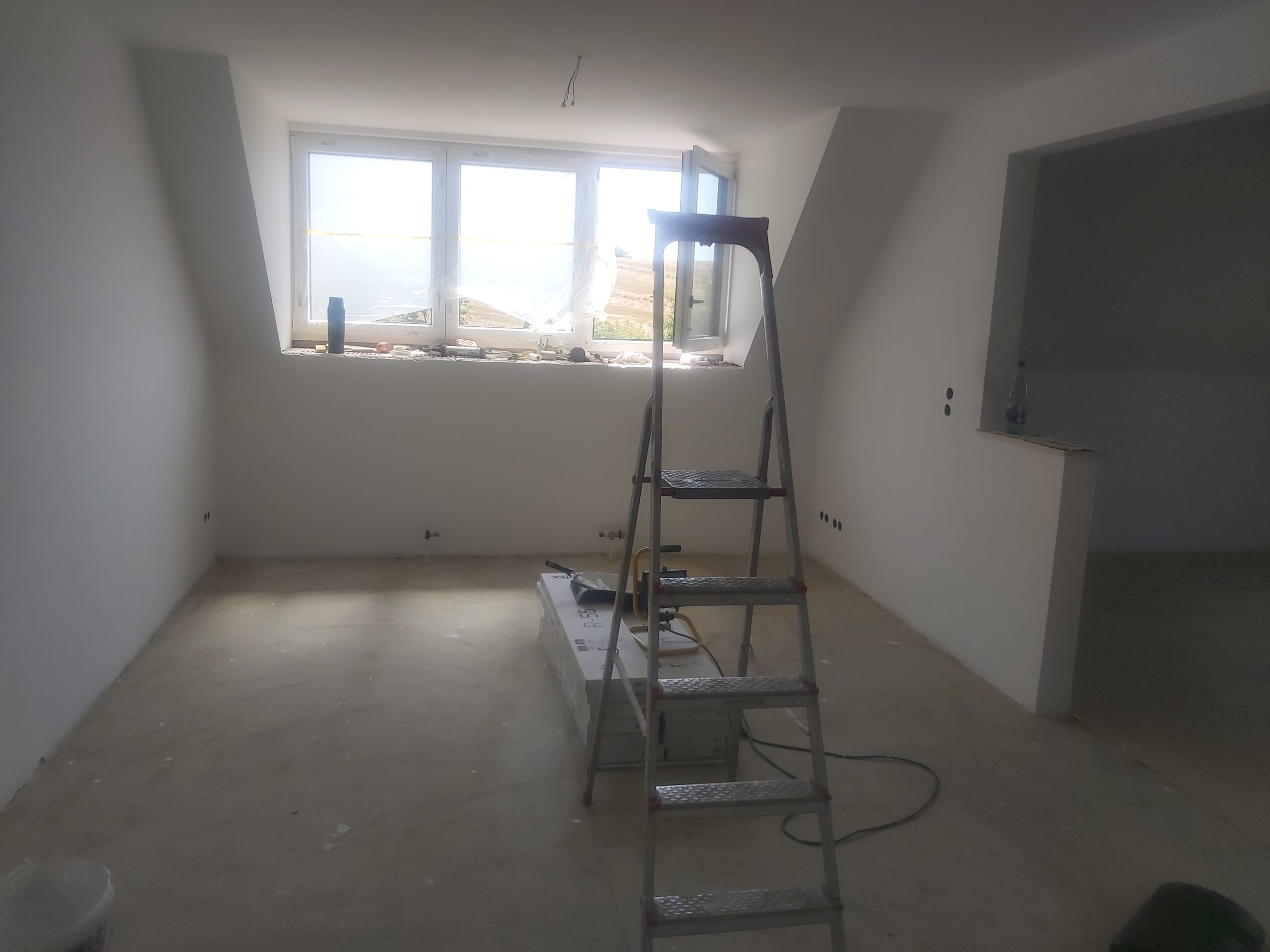 Pokój na poddaszu w stanie surowym po pracach wykończeniowych, z białymi ścianami, oknem dachowym, drabiną stojącą na środku oraz widocznymi narzędziami i przewodami na podłodze.