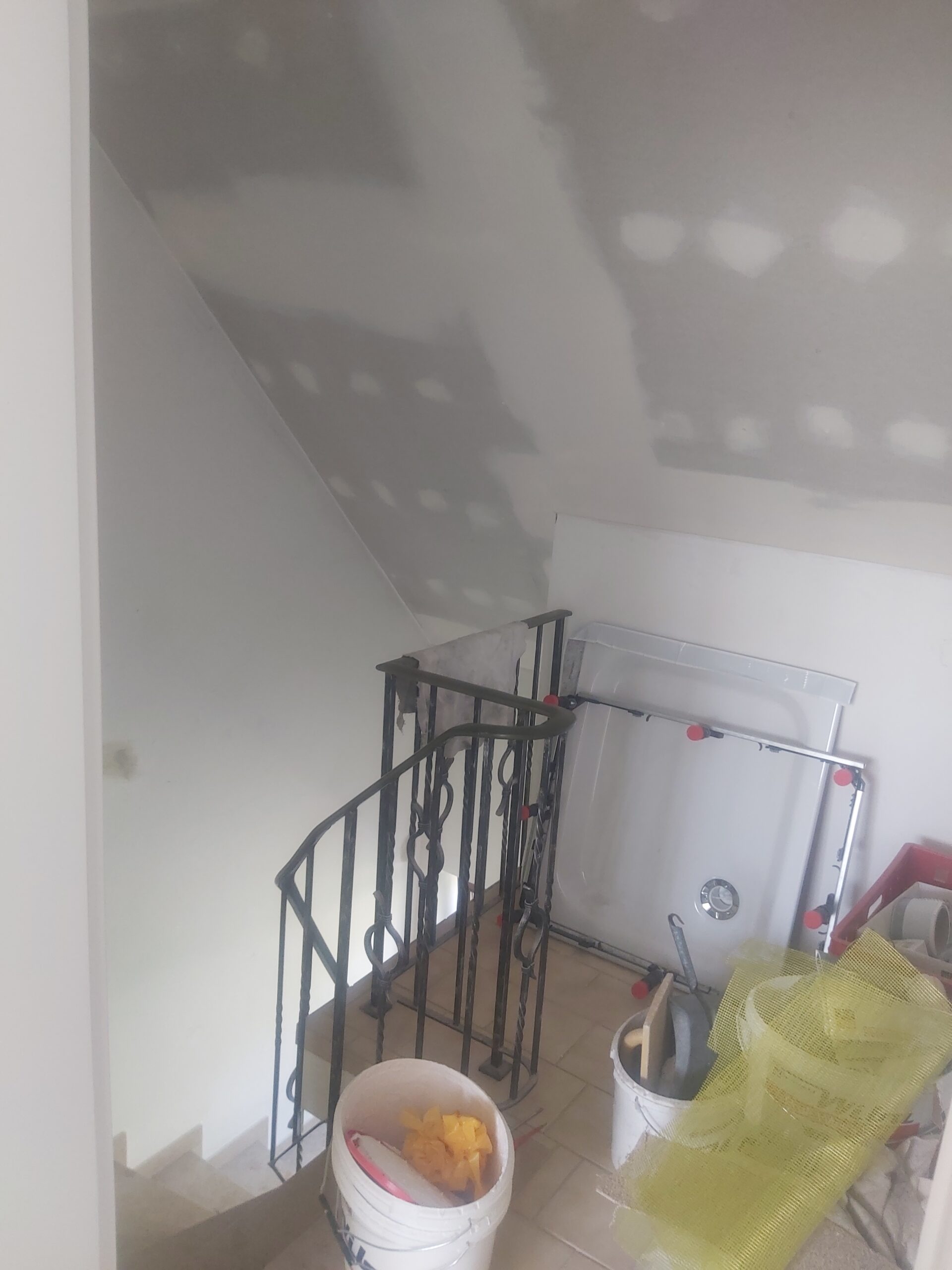 Klatka schodowa na poddaszu podczas remontu, z widoczną balustradą, wiadrami z narzędziami i materiałami budowlanymi oraz sufitem z płyt gipsowych przygotowanym do wykończenia.