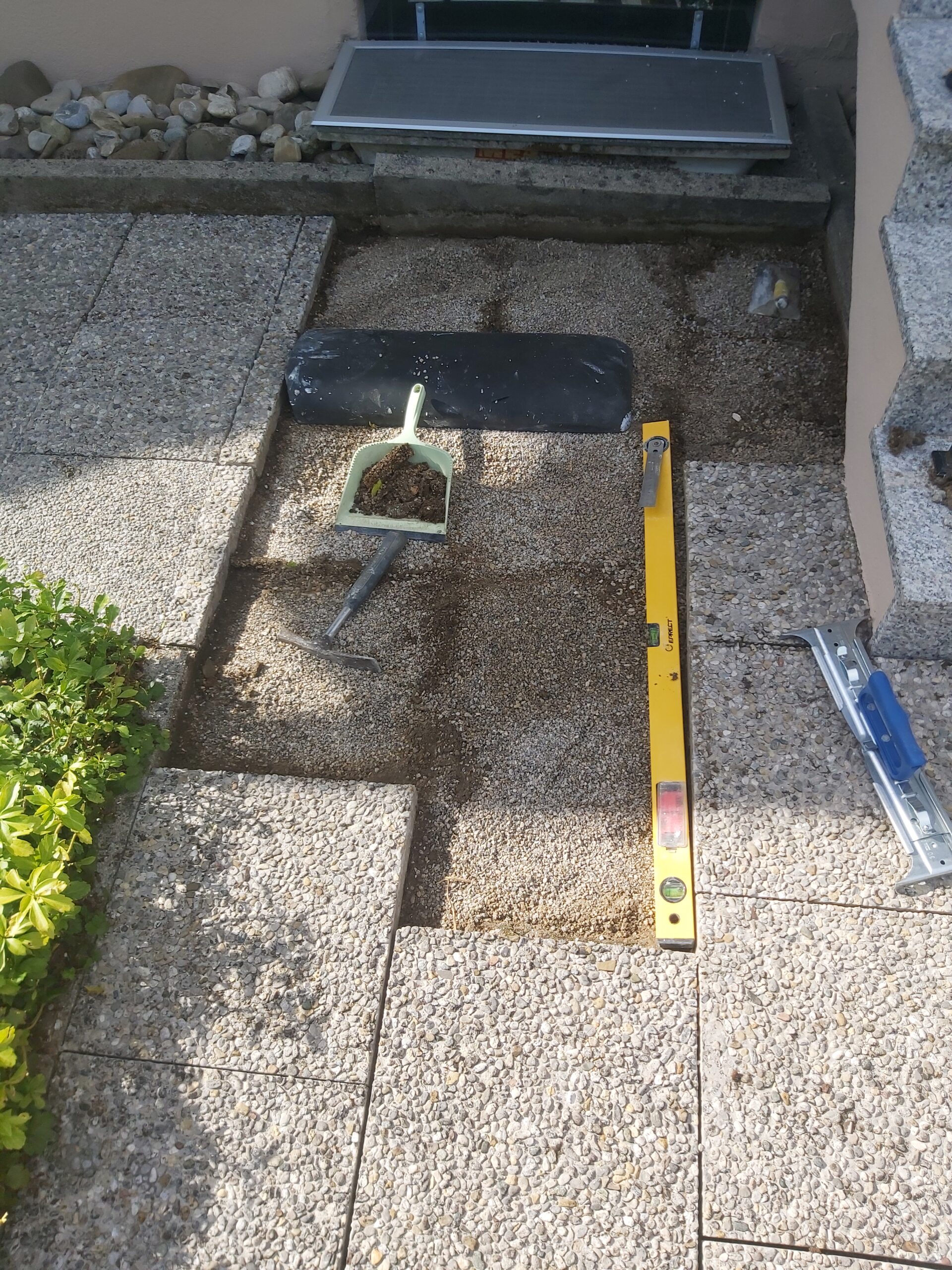 Fragment przygotowanego podłoża z podsypką pod kostkę brukową przy wejściu do budynku, z widoczną poziomicą i narzędziami używanymi podczas układania nawierzchni.