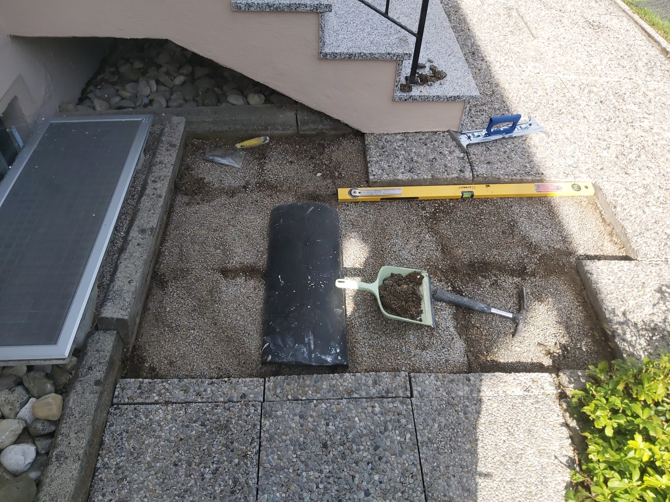 Przygotowany fragment nawierzchni z kostki brukowej przy schodach z widocznymi narzędziami brukarskimi, poziomicą i szuflą podczas prac wykończeniowych.