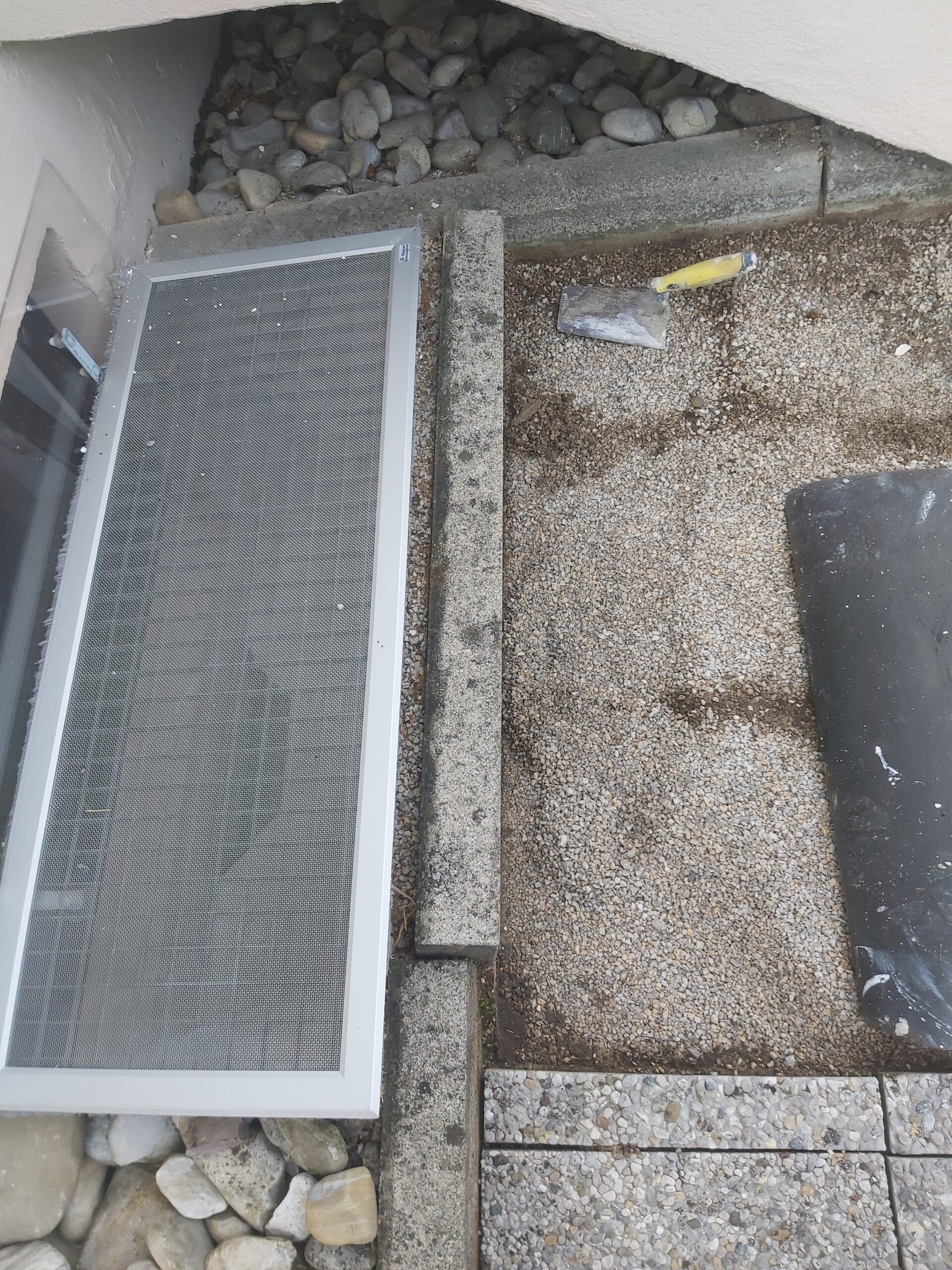 Odsłonięte podłoże pod płyty chodnikowe przy wejściu do budynku, z widoczną kratką okienną piwnicy, obrzeżem betonowym oraz narzędziami do prac brukarskich.