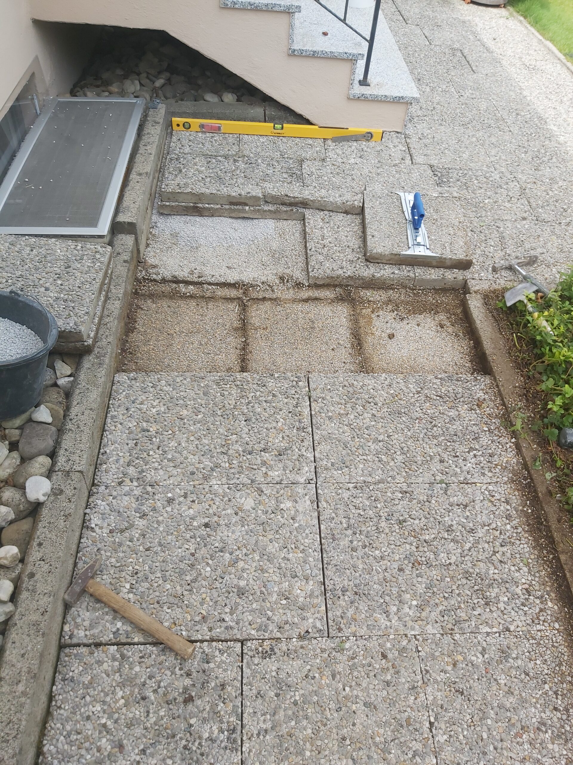 Fragment kamiennego chodnika zdemontowany przed schodami wejściowymi, z widoczną poziomicą, narzędziami oraz odsłoniętą podsypką pod płytami.
