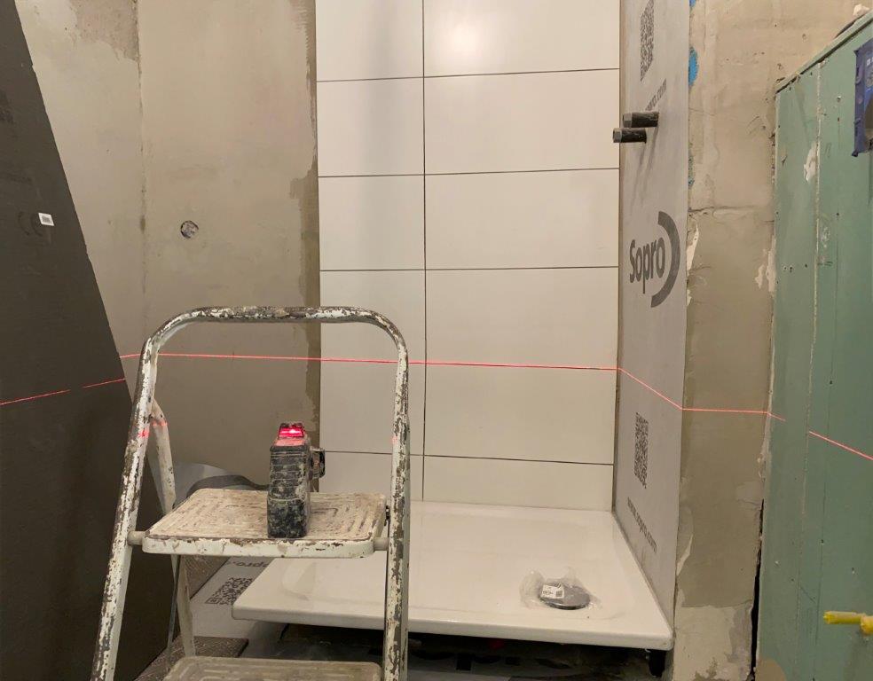 Układanie białych płytek ściennych w strefie prysznica, widoczny laser poziomujący, brodzik oraz surowe ściany przygotowane do dalszych prac.