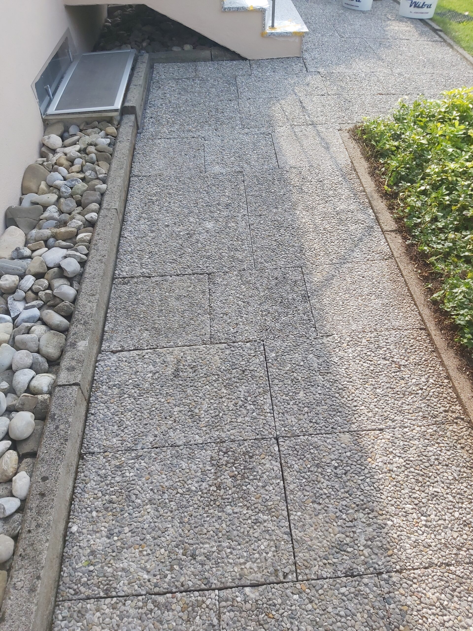 Kamienny chodnik prowadzący wzdłuż elewacji domu, z widocznym obrzeżem, ozdobnymi kamieniami po jednej stronie i zielonym żywopłotem po drugiej.