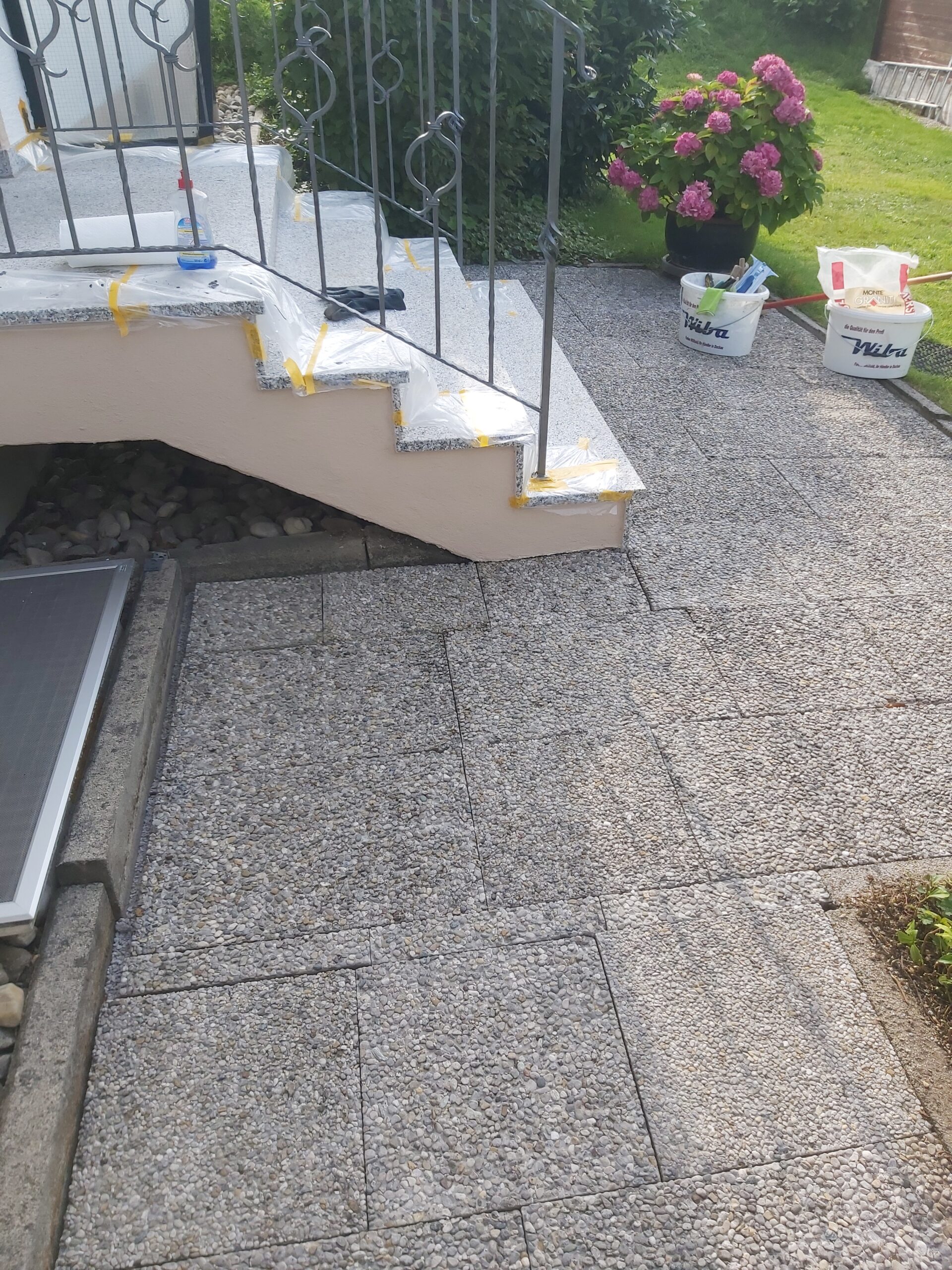 Zewnętrzne schody prowadzące do wejścia do domu, zabezpieczone folią i taśmą malarską, z widocznym kamiennym tarasem oraz wiadrami z materiałami budowlanymi obok.