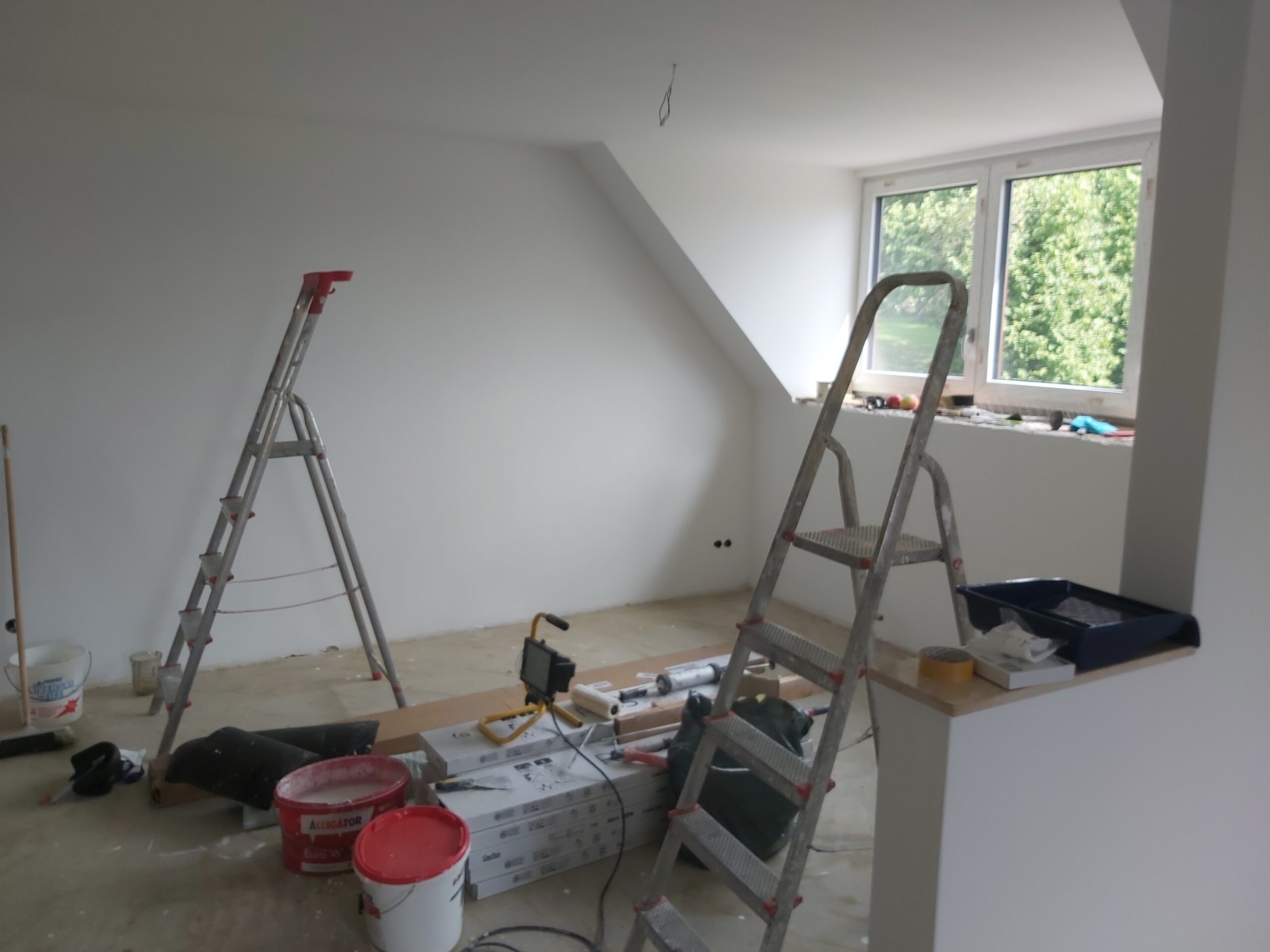 Pomieszczenie na poddaszu w trakcie remontu z widocznymi drabinami, narzędziami budowlanymi, materiałami wykończeniowymi oraz oknem doświetlającym wnętrze.