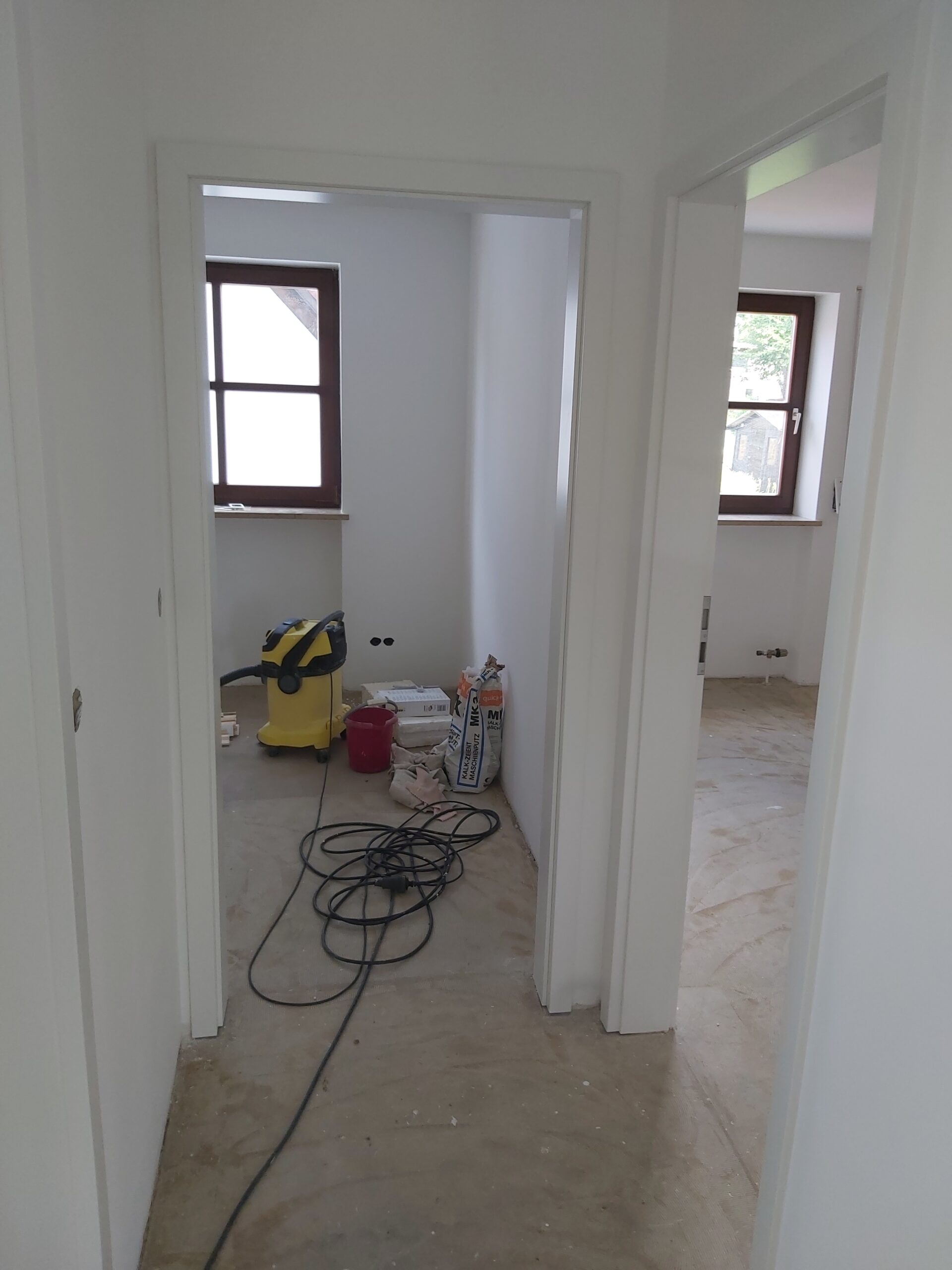 Korytarz w trakcie remontu z widocznymi wejściami do dwóch pomieszczeń, narzędziami budowlanymi na podłodze oraz nieukończonymi pracami wykończeniowymi.