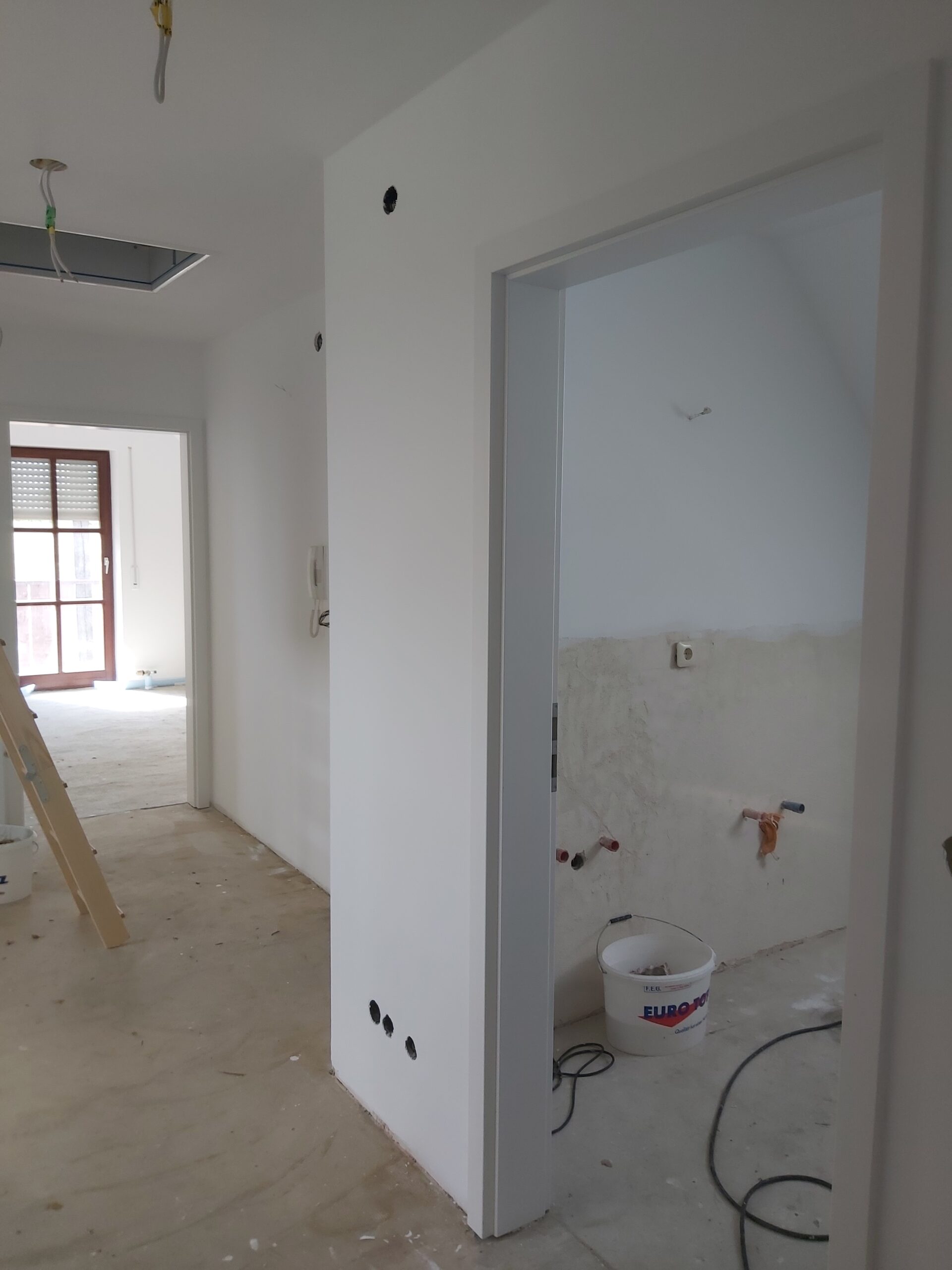 Korytarz w trakcie remontu z widocznym wejściem do łazienki, nieukończonymi instalacjami elektrycznymi, przygotowanymi ścianami oraz narzędziami budowlanymi.