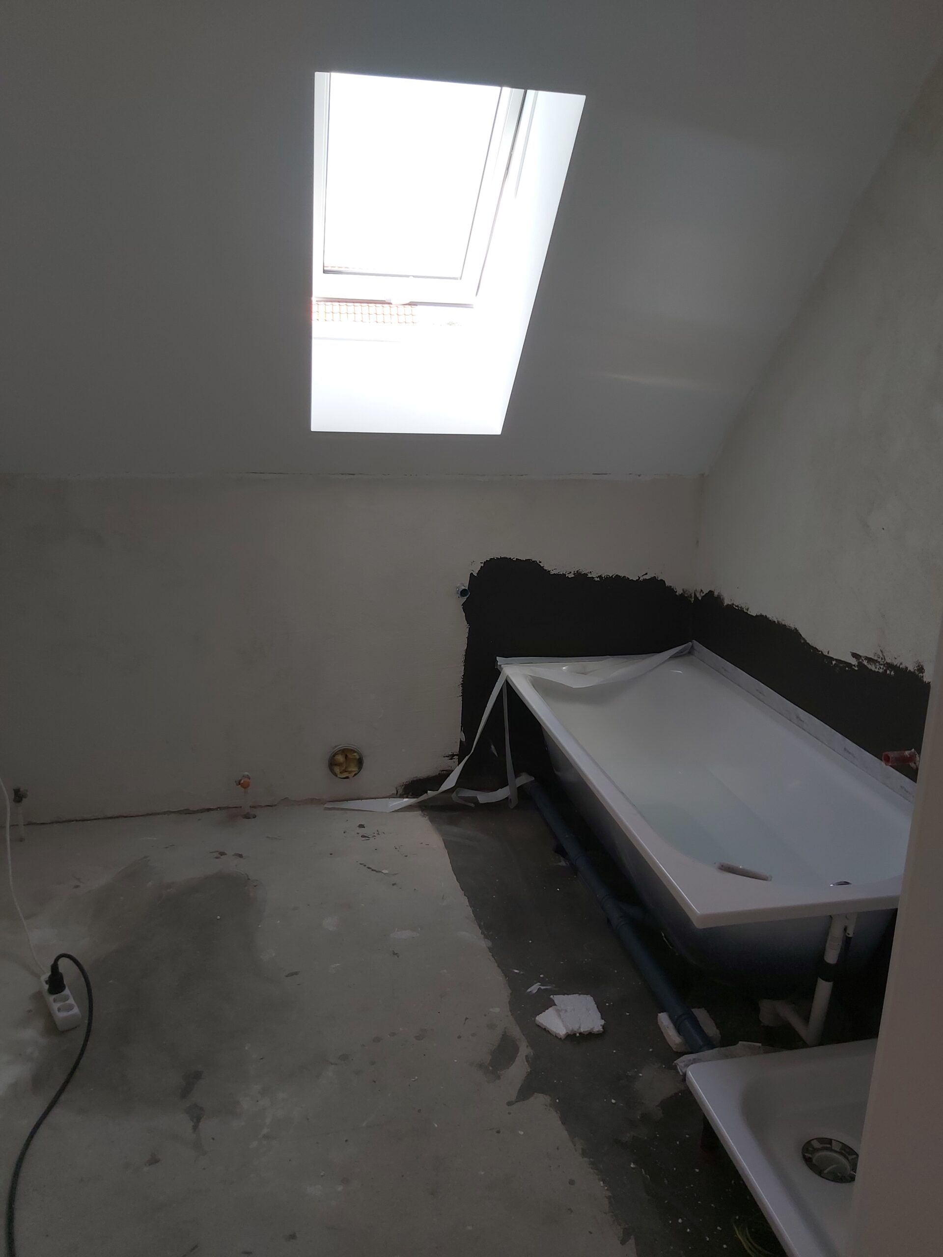 Łazienka na poddaszu w trakcie prac wykończeniowych z zamontowaną wanną, widoczną izolacją ściany, oknem dachowym oraz nieukończoną posadzką.