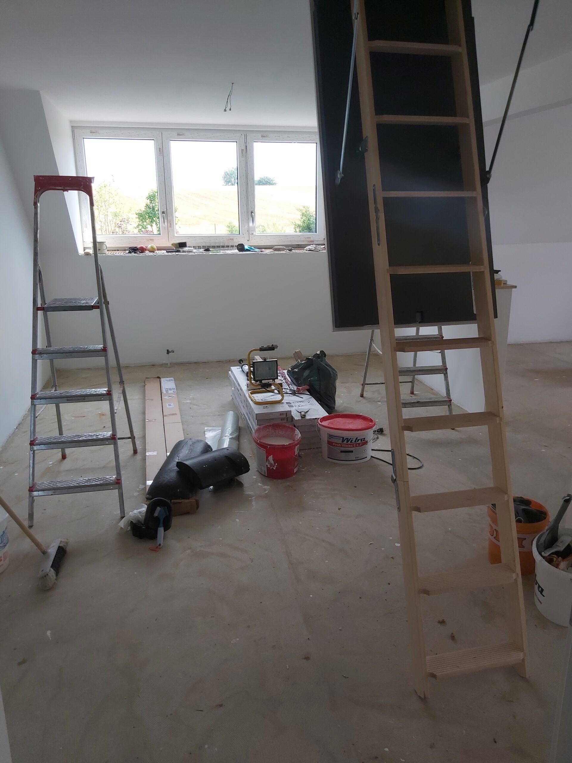 Wnętrze poddasza w trakcie remontu z rozstawionymi drabinami, narzędziami budowlanymi, wiadrami z materiałami oraz oknami dachowymi wpuszczającymi światło dzienne.