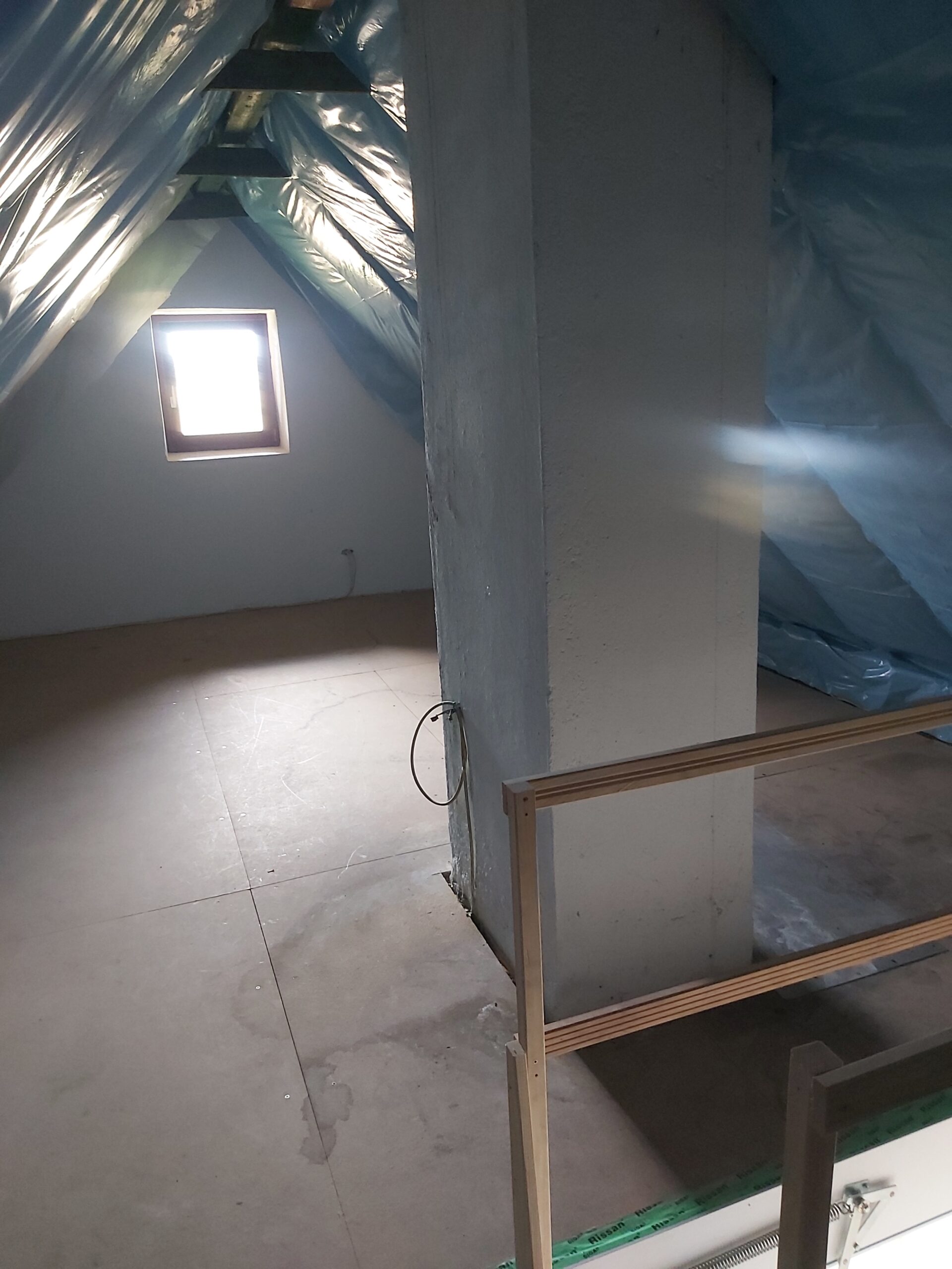 Poddasze w trakcie prac wykończeniowych z widocznym kominem, folią paroizolacyjną pod połaciami dachowymi oraz ułożoną podłogą z płyt.